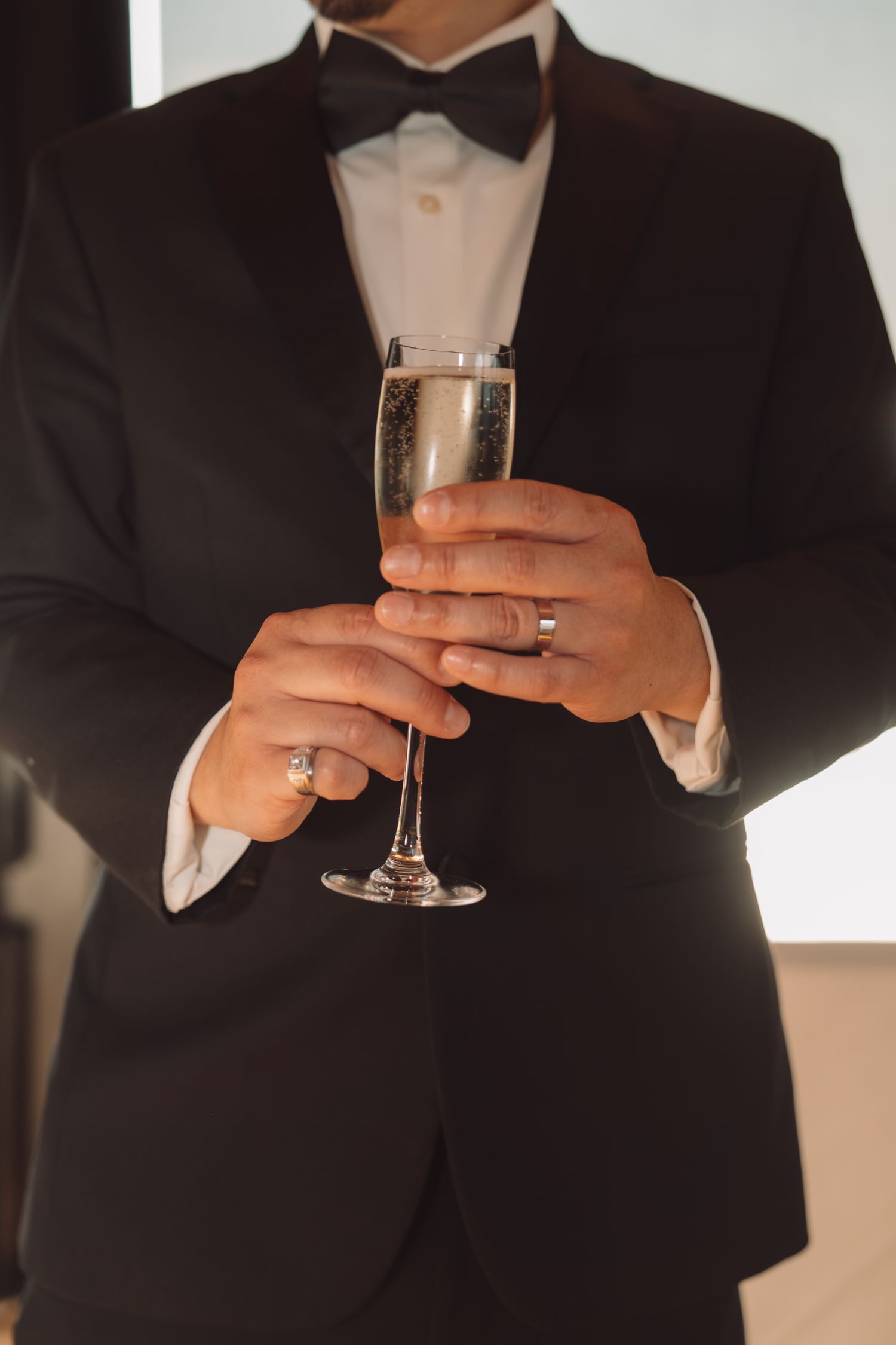 Groom in black tuxedo with bow tie holding a champagne flute, wearing a wedding ring at a formal celebration.