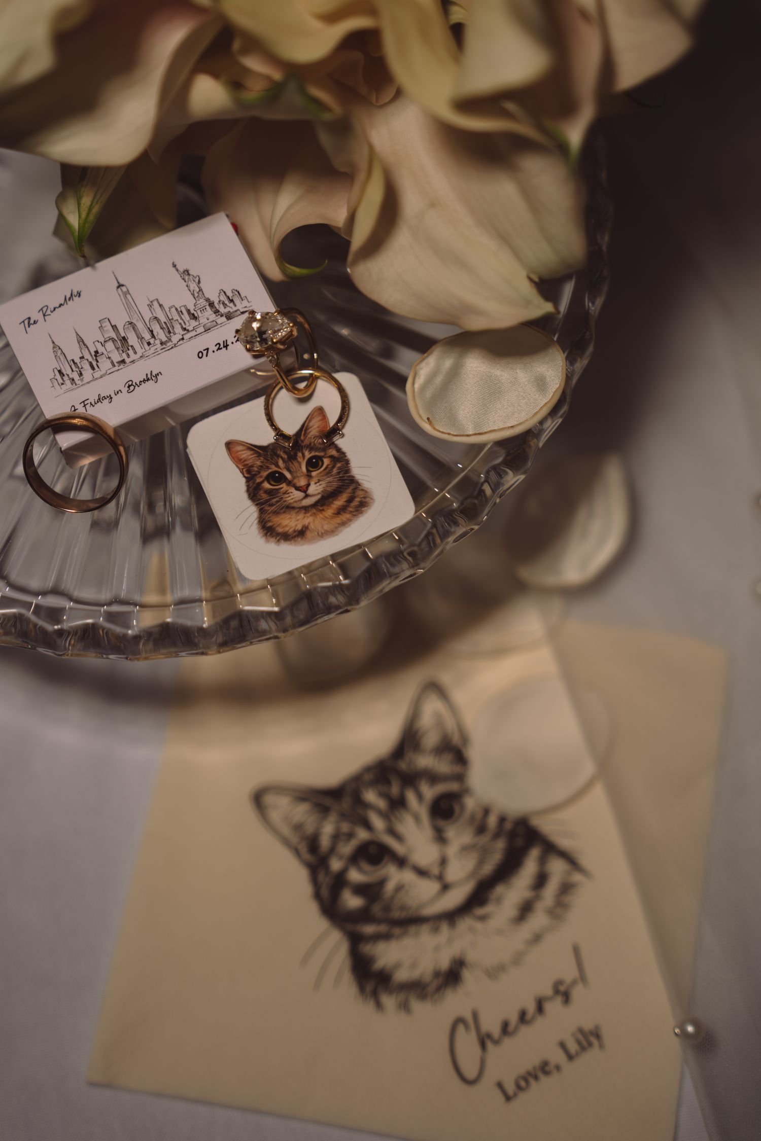 Wedding flat lay with cat keychain, rings on crystal dish, and illustrated cat card reading Cheers Love Lily.