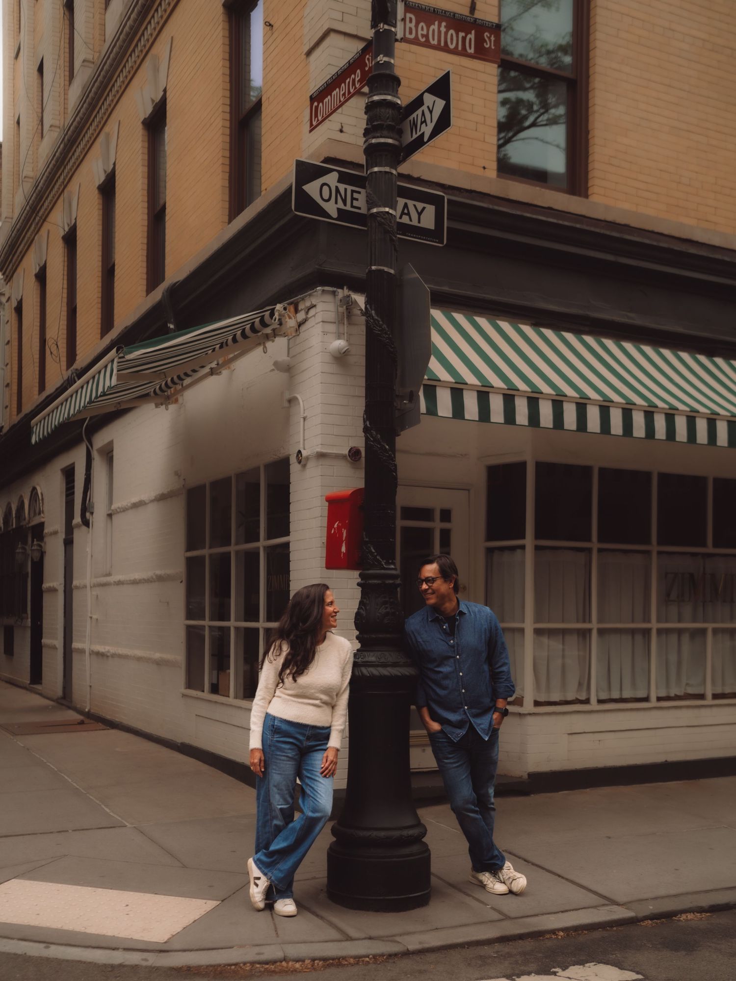 Couple leaning against a street pole at Bedford St corner in NYC, smiling at each other on a quiet urban sidewalk.