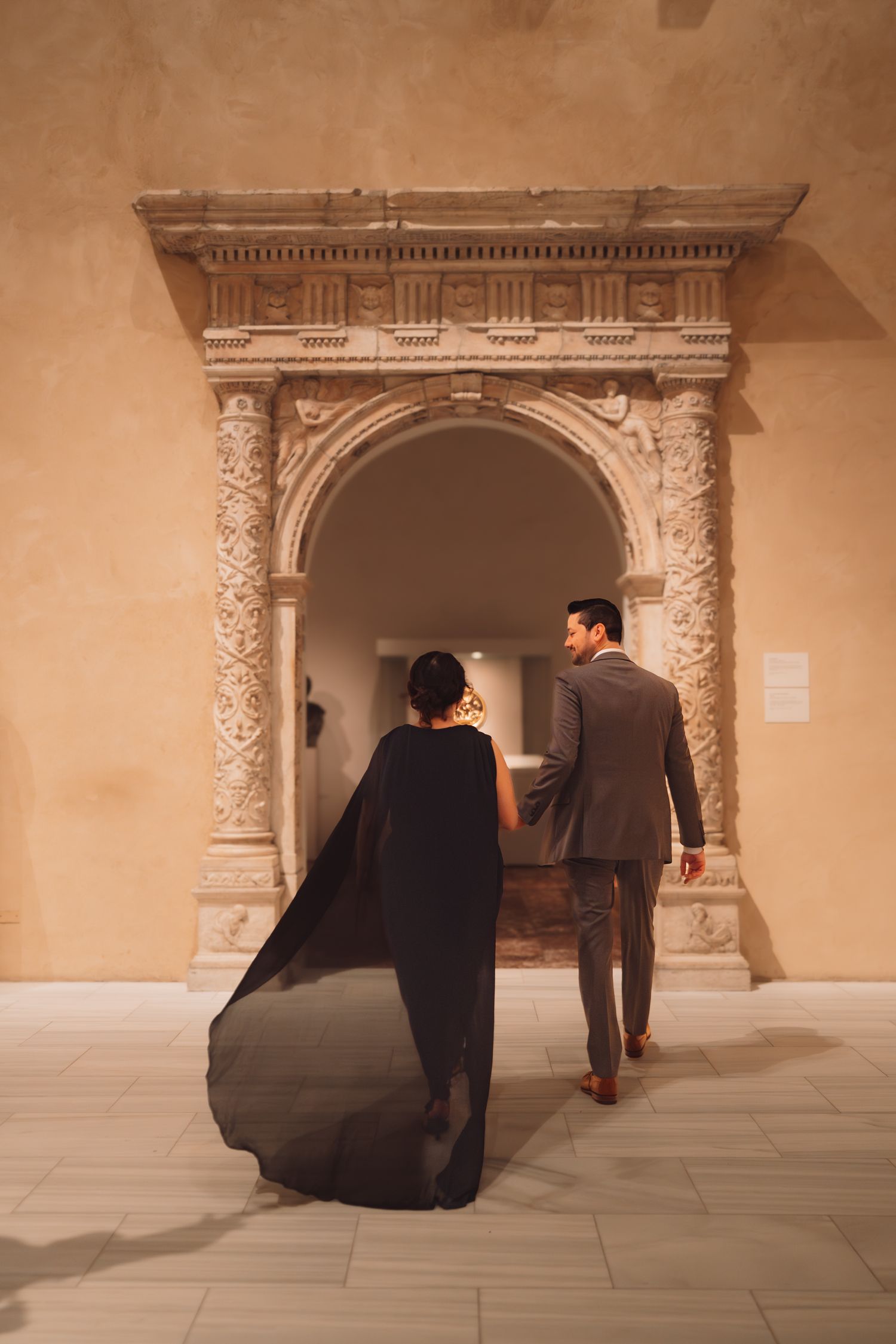 Elegant couple in formal attire walking through ornate stone archway in a museum gallery.