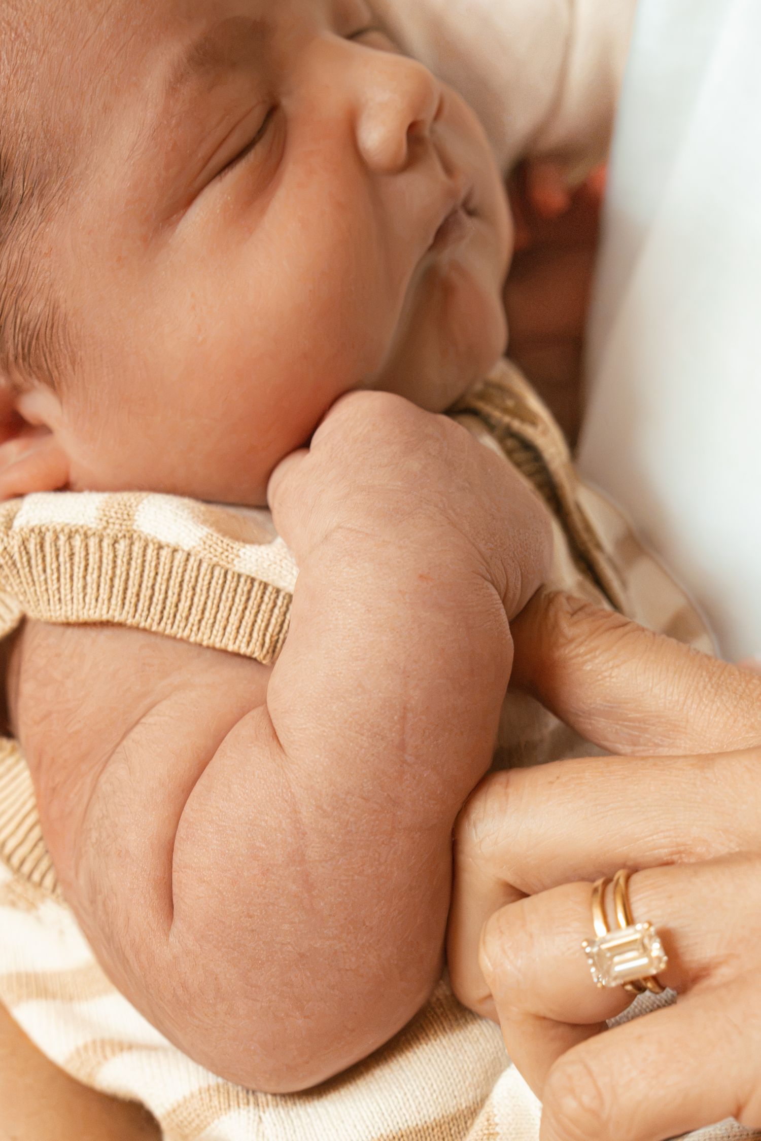 Sleeping newborn baby cradled in mother's arms, wearing a knit outfit, with mother's gold ring visible.