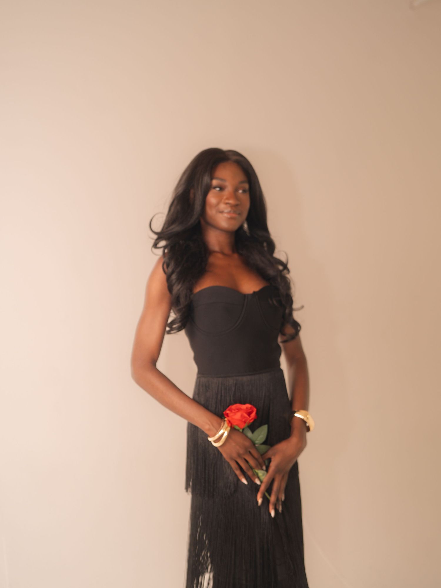 Young woman in black strapless fringe dress holding a red rose, wearing gold bracelets, posing against a neutral background.