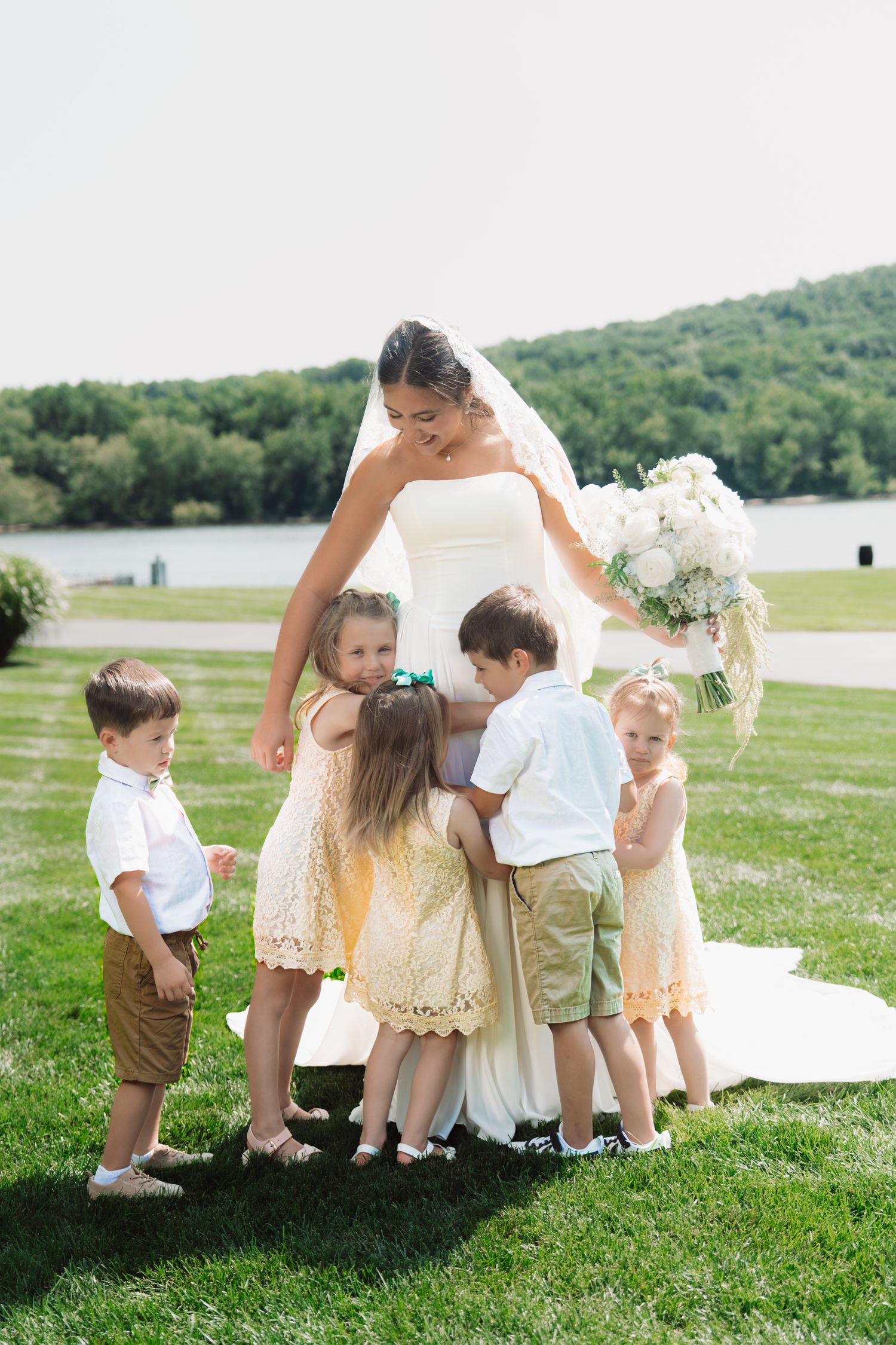 Bride in white gown holds bouquet while flower girls and ring bearers hug her on green lawn with lake backdrop.