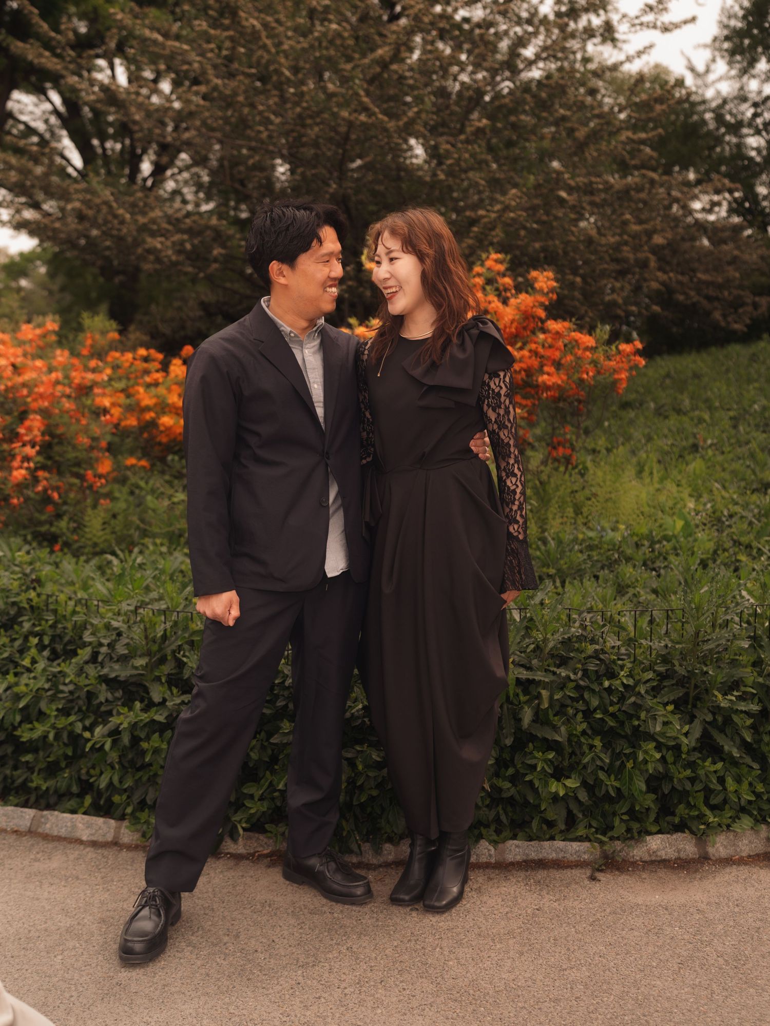 A couple smiles at each other in a park with orange flowers and green trees in the background.