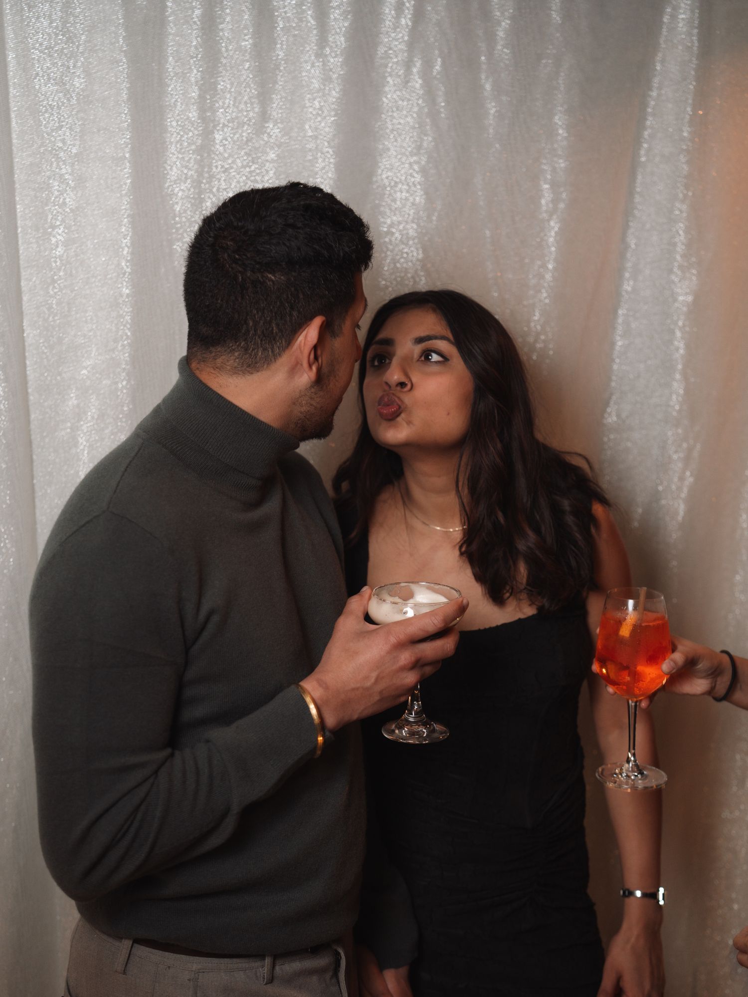 Couple posing playfully with cocktails at a glittery white sequin photo booth backdrop.