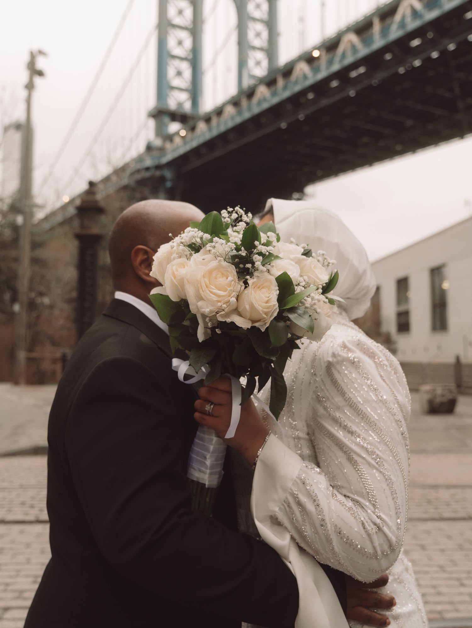 Couple sharing a kiss behind a white rose bouquet under the Manhattan Bridge in Brooklyn, NYC.