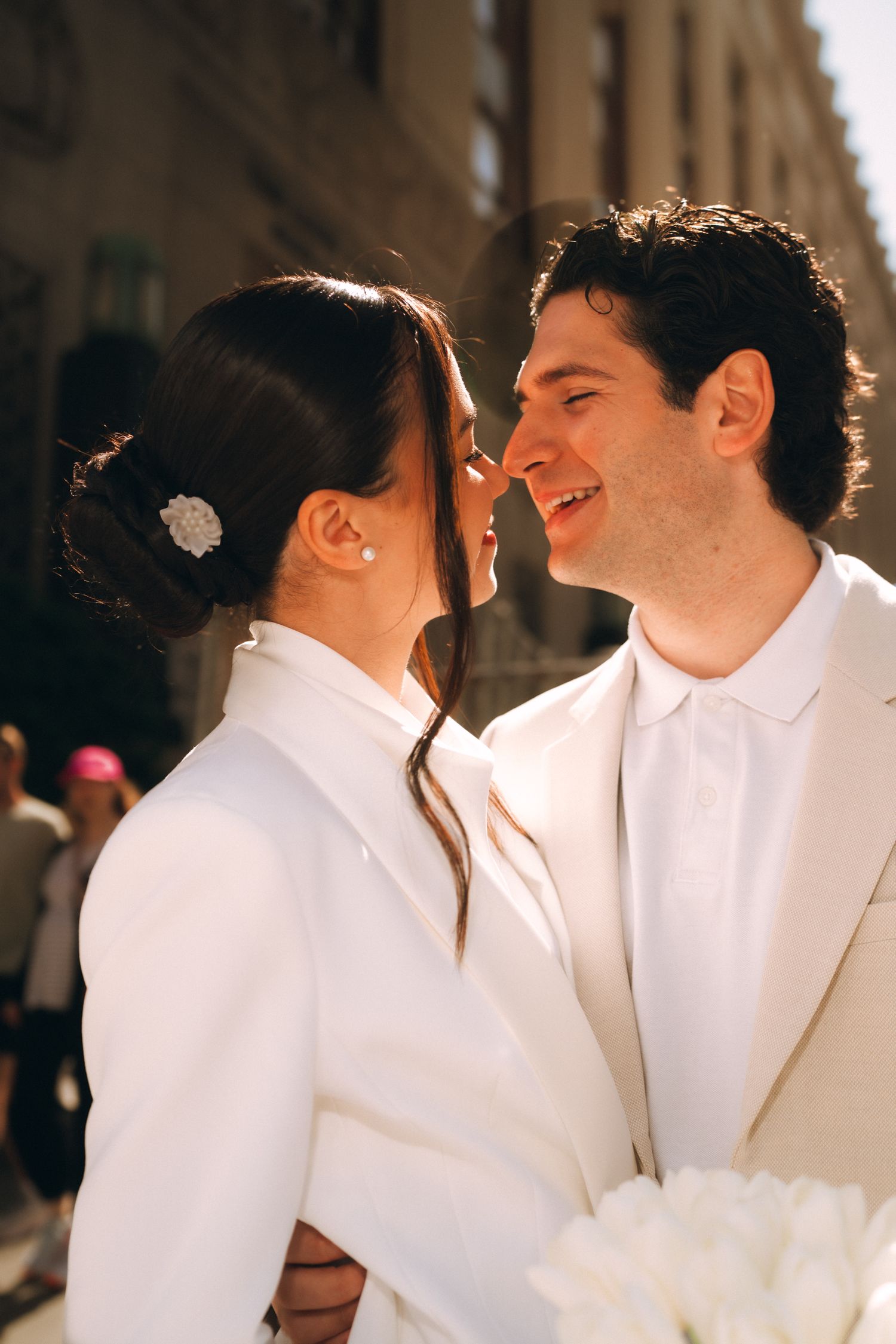 Bride and groom in matching white suits share a romantic moment on a sunny city street.