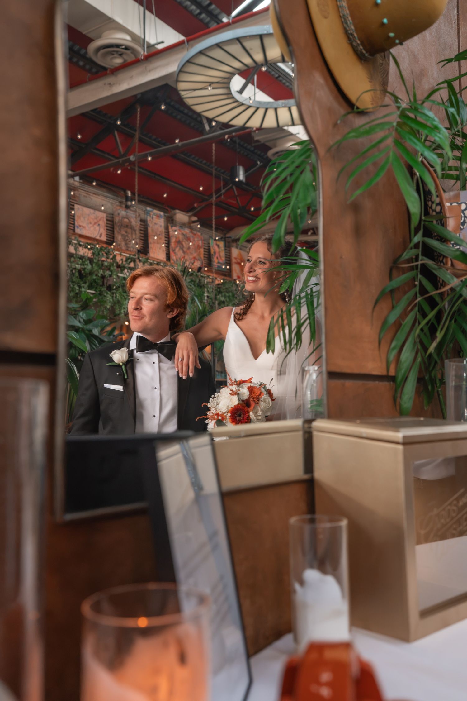 Couple in wedding attire reflected in mirror, bride holding bouquet, groom in tuxedo, surrounded by lush greenery and warm lights.