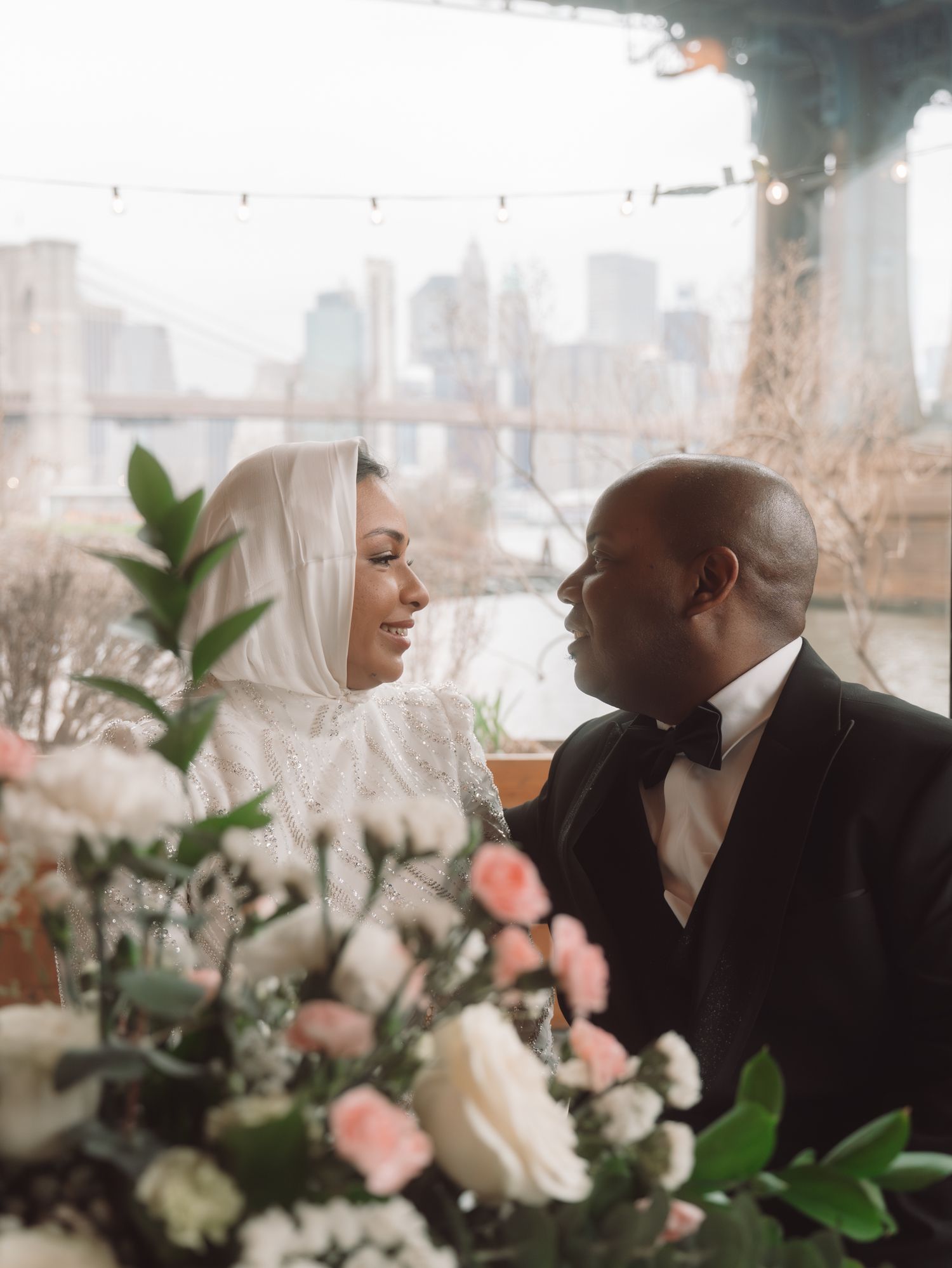 Muslim bride in white hijab and groom gaze lovingly at each other near Brooklyn Bridge.