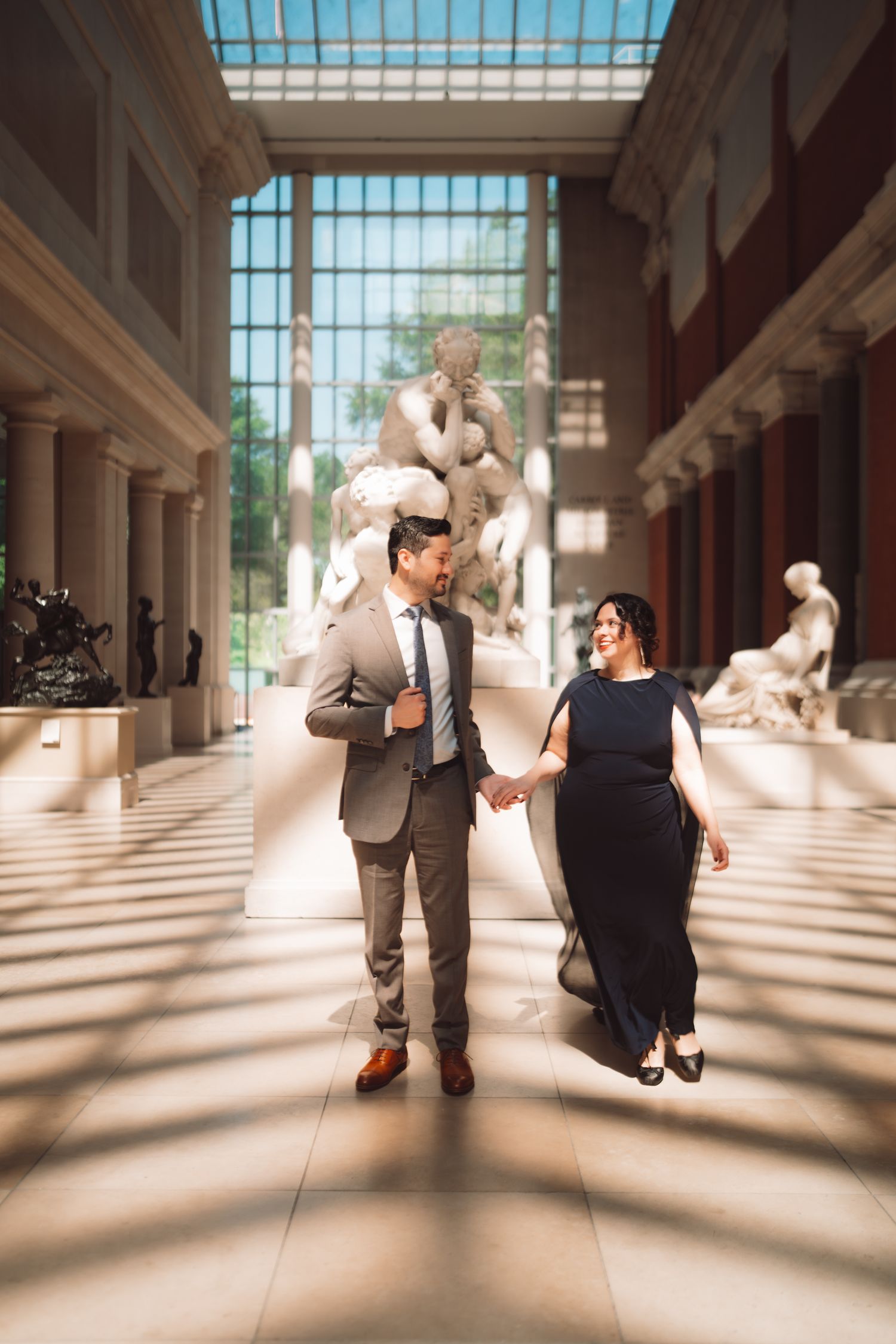 Couple holding hands walking through a grand museum gallery with classical sculptures and natural light.