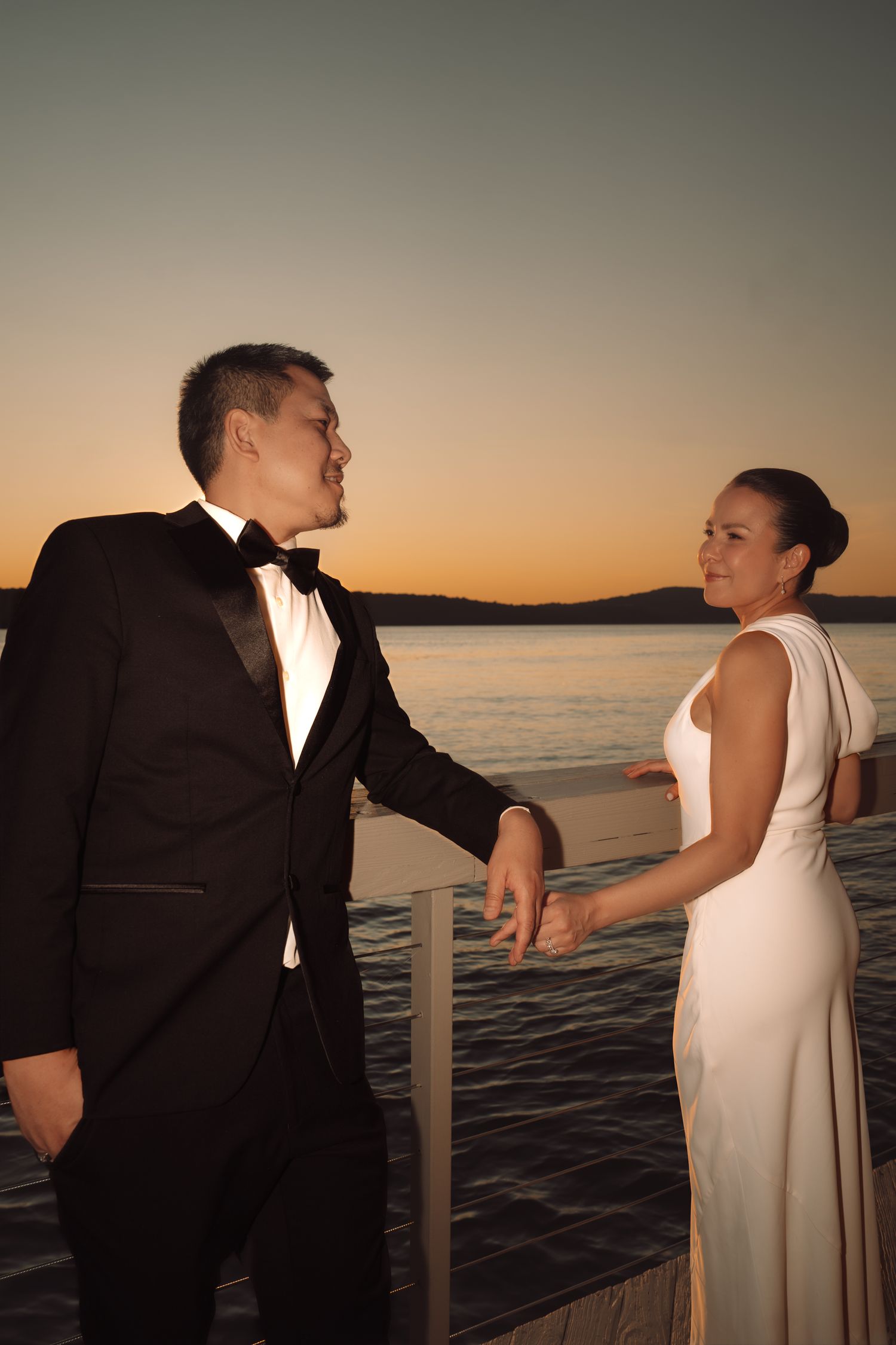 Couple in formal attire holding hands on a waterfront dock at sunset, man in tuxedo and woman in white gown.