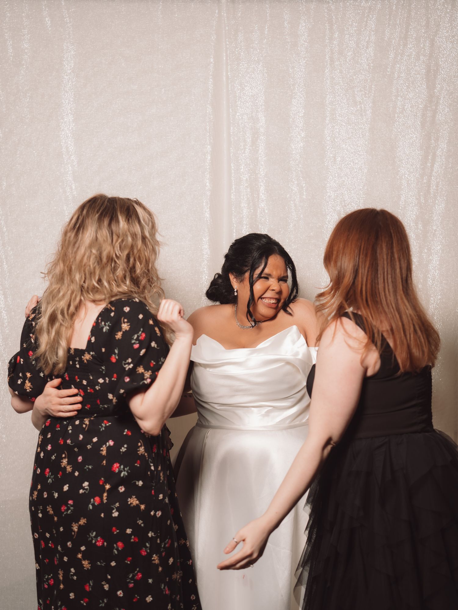 Bride in white satin gown laughing joyfully with two female guests at wedding reception.