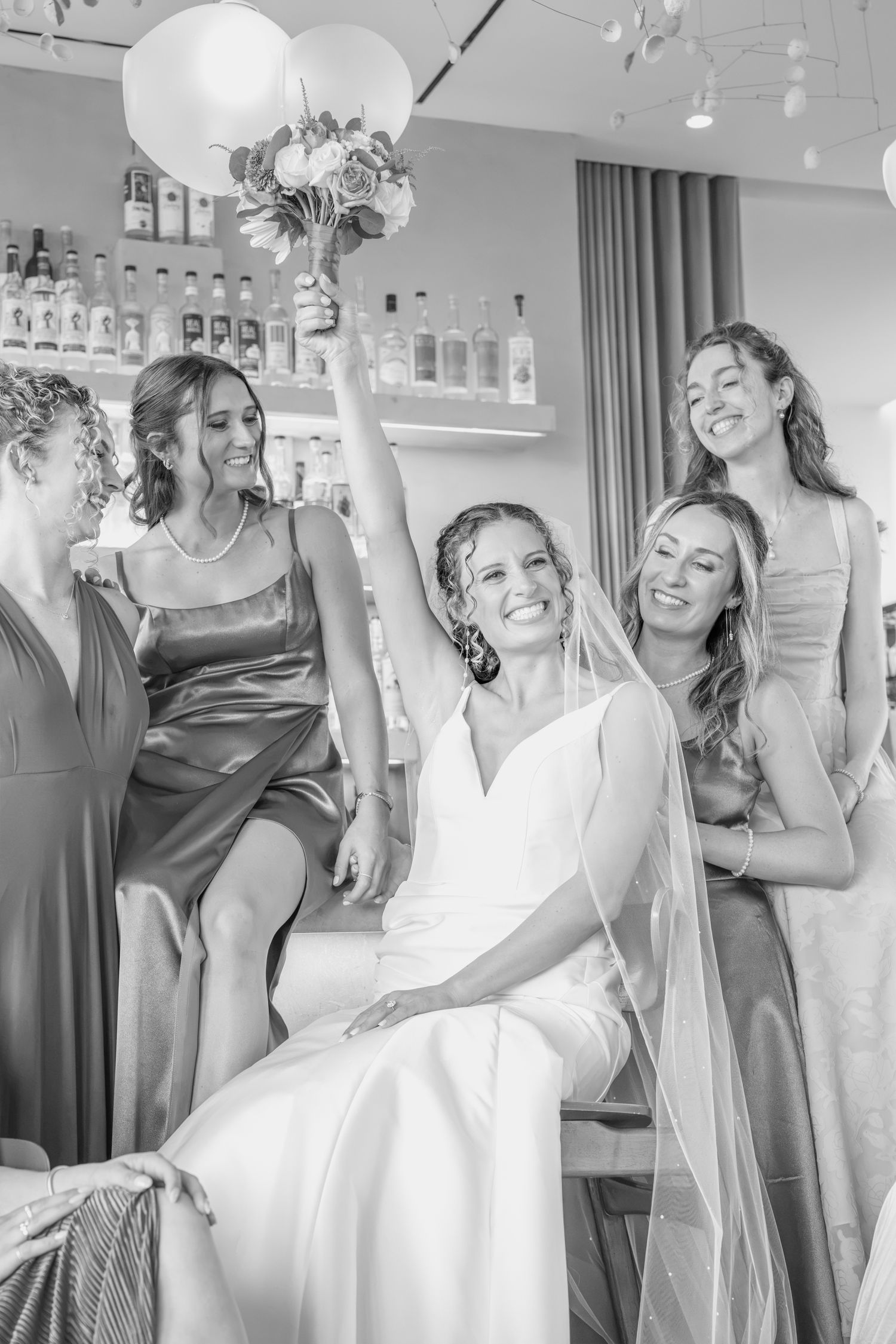 Bride in white gown raises bouquet surrounded by smiling bridesmaids in satin dresses near a bar.