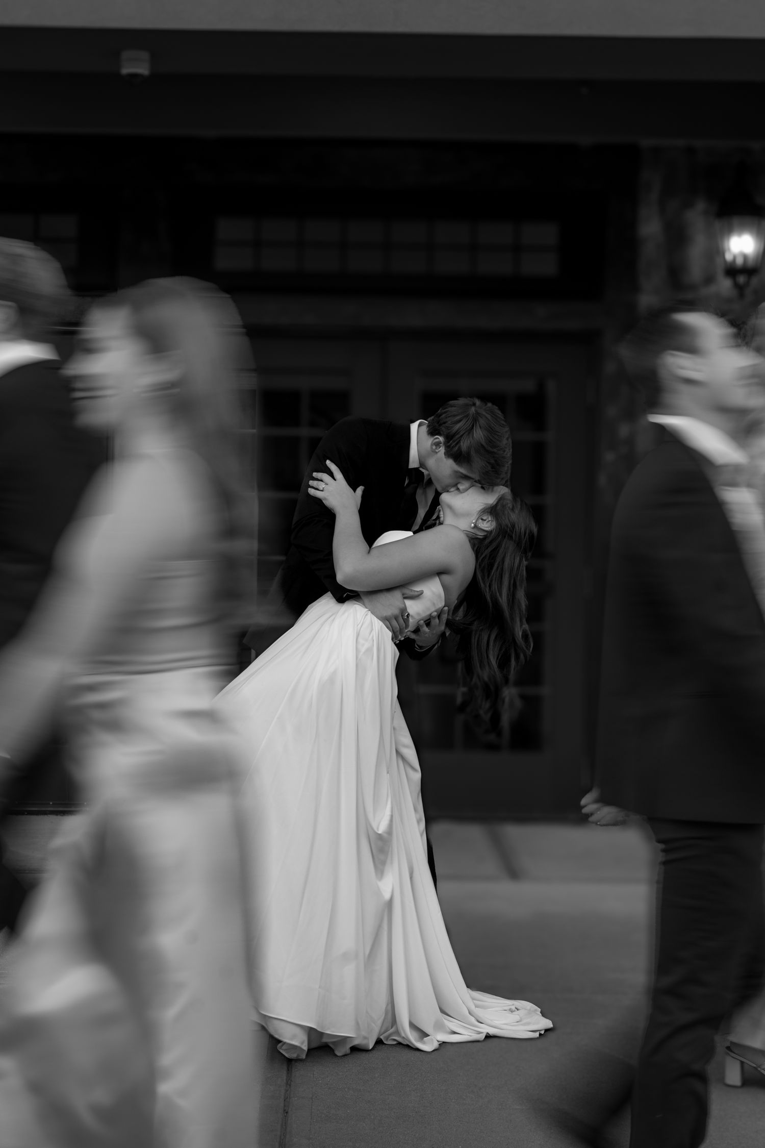 Black and white photo of a couple embracing and kissing in a white gown amid blurred moving guests.