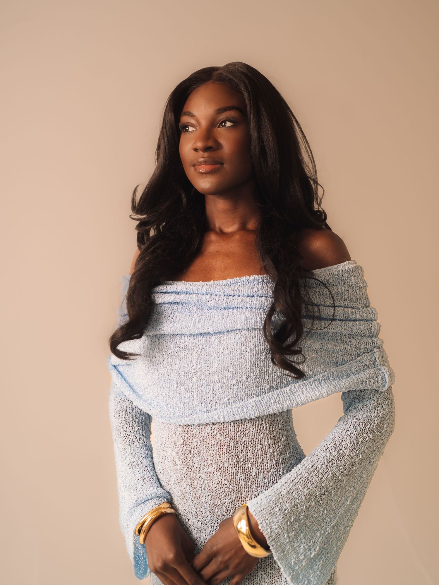 Young woman in light blue off-shoulder knit dress with gold bangles posing against beige background.