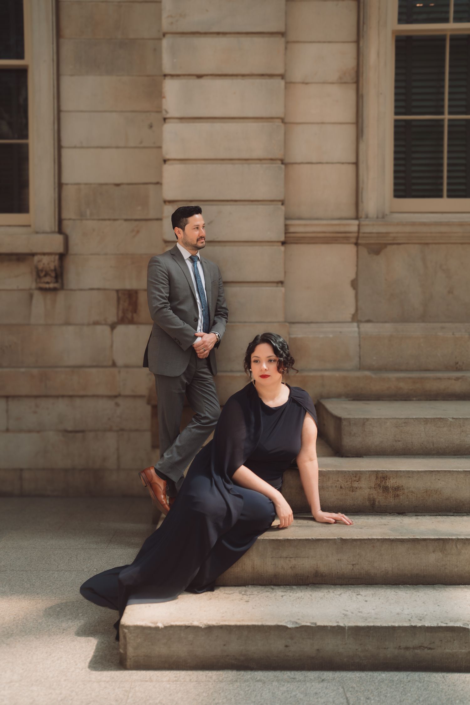 A woman in a black gown sits on stone steps while a man in a gray suit stands behind her outside a building.