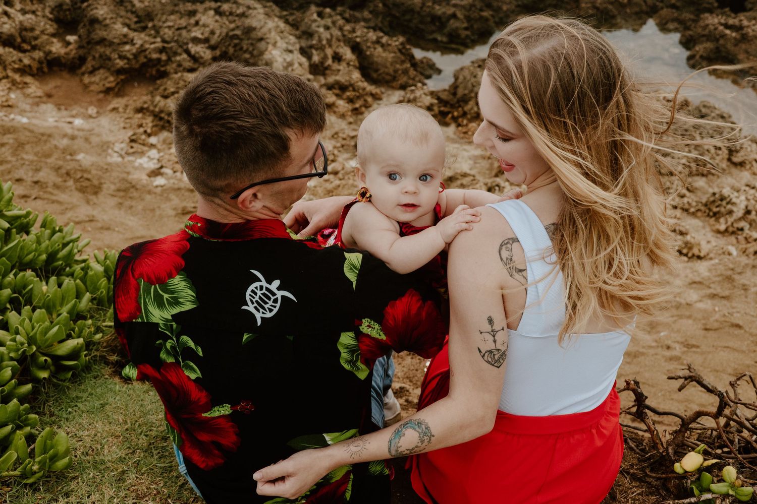 Parents in red floral and white outfits holding baby near rocky beach shoreline.