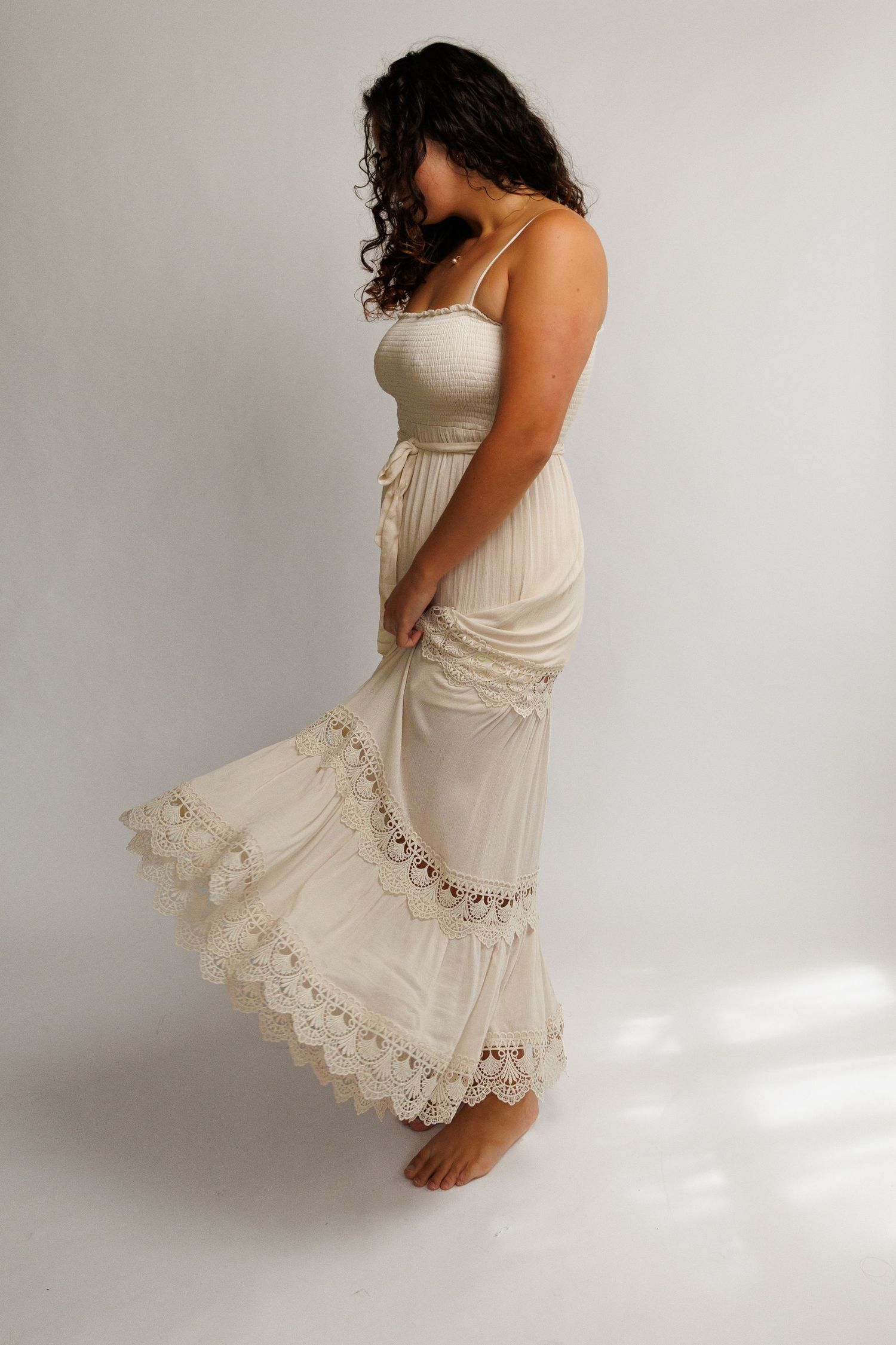 Woman in cream boho maxi dress with lace trim and smocked bodice, flowing skirt, barefoot on white background.