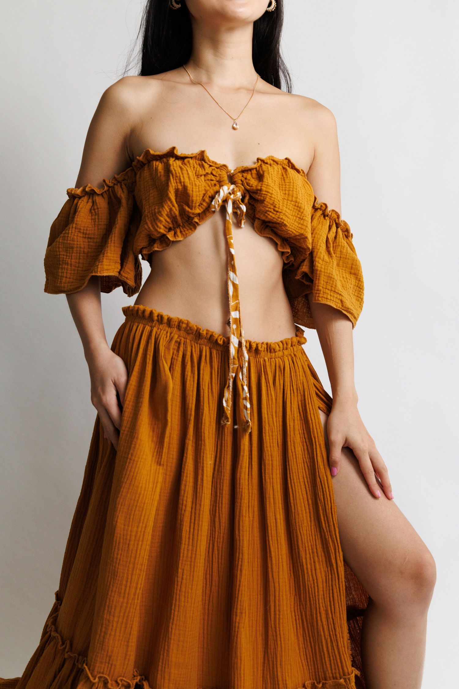 Woman wearing a mustard yellow off-shoulder crop top and maxi skirt set with ruffle details and side slit.