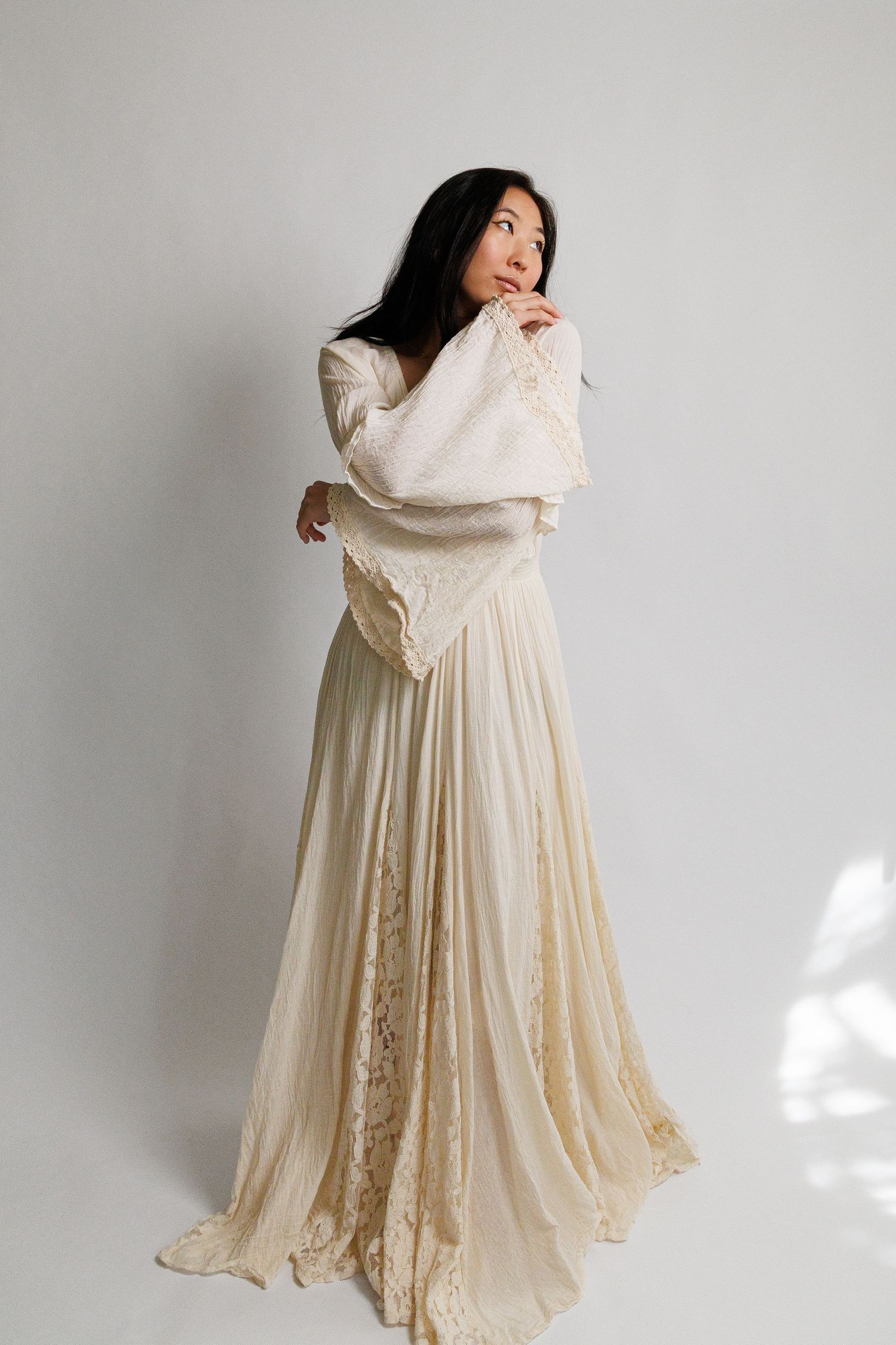 Woman in elegant ivory lace wedding gown with long sleeves and flowing skirt, posing thoughtfully in studio.