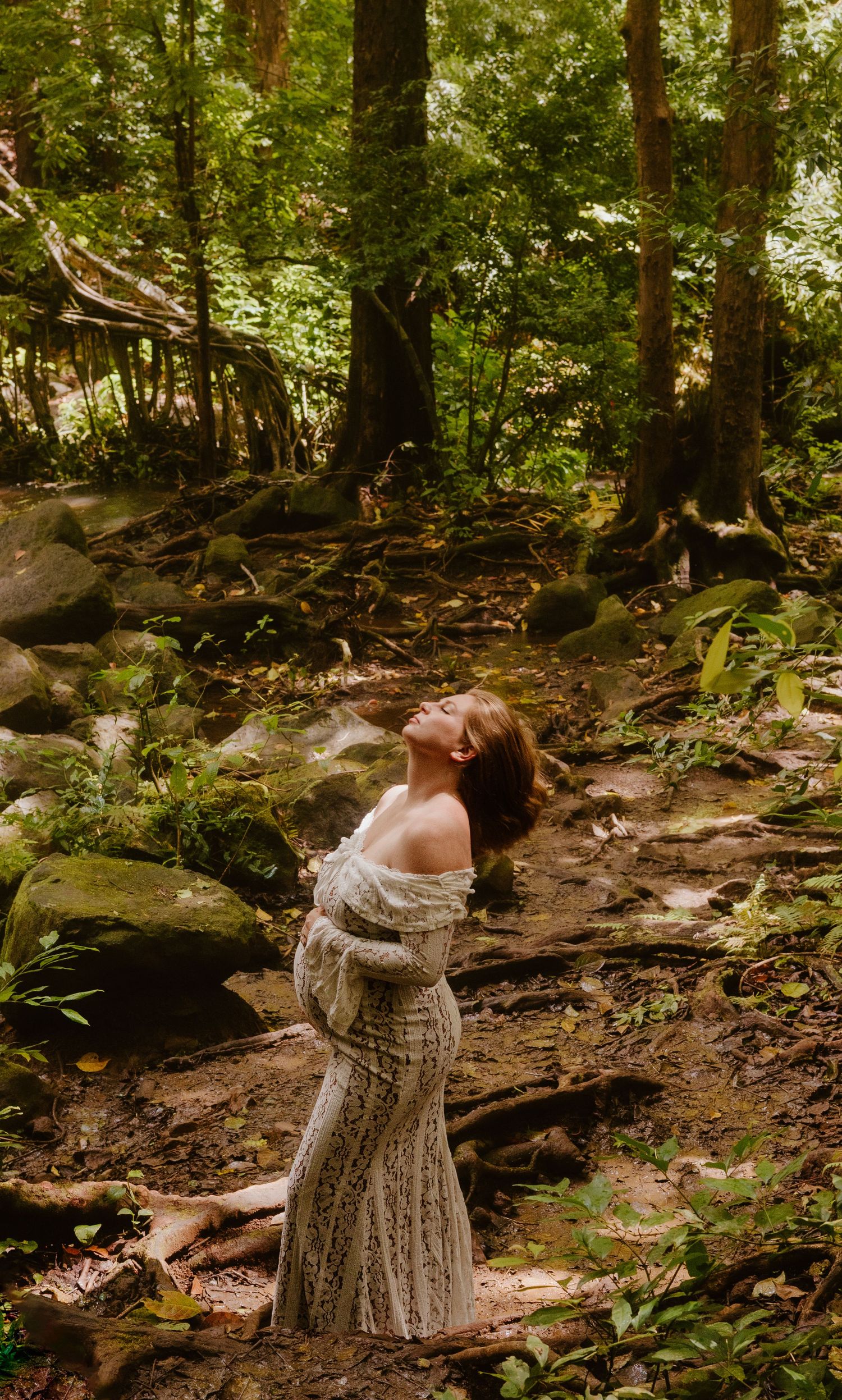 Pregnant woman in off-shoulder lace gown standing in lush green forest with head tilted back.
