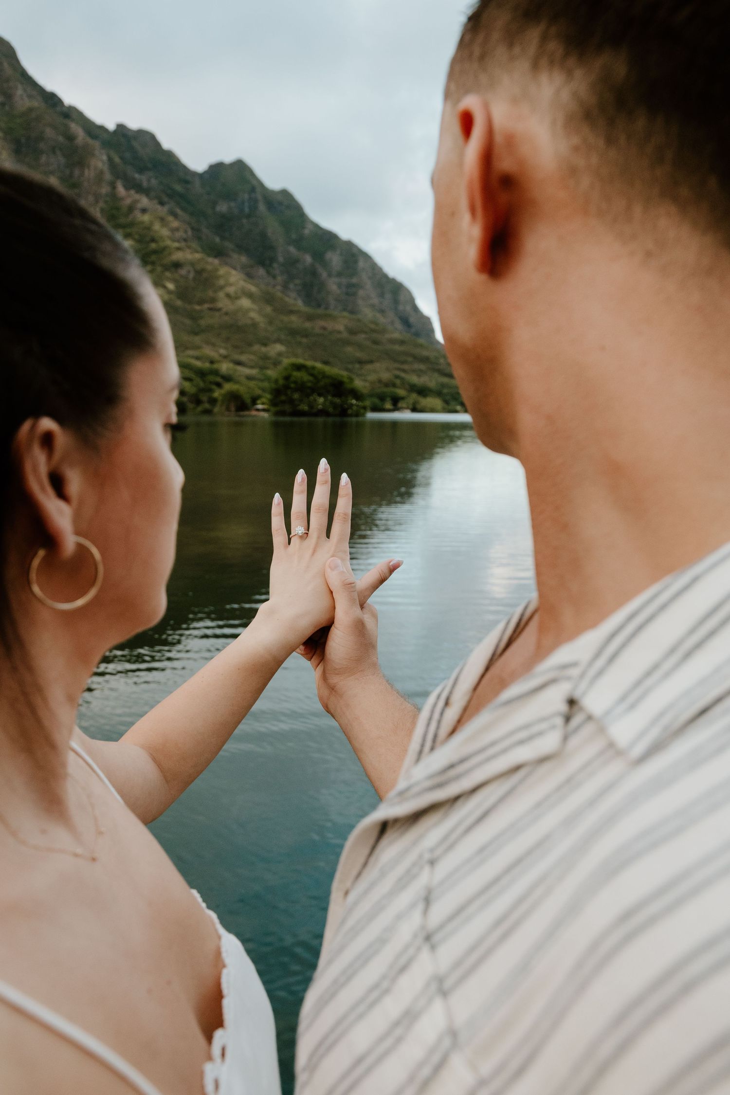 Engaged couple admiring diamond ring together on dock with scenic mountain lake in background.