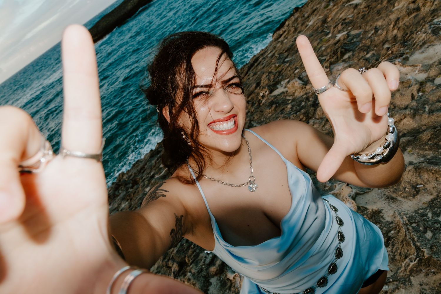 Smiling woman making finger gun gestures at camera near rocky ocean shore, wearing blue top and jewelry.