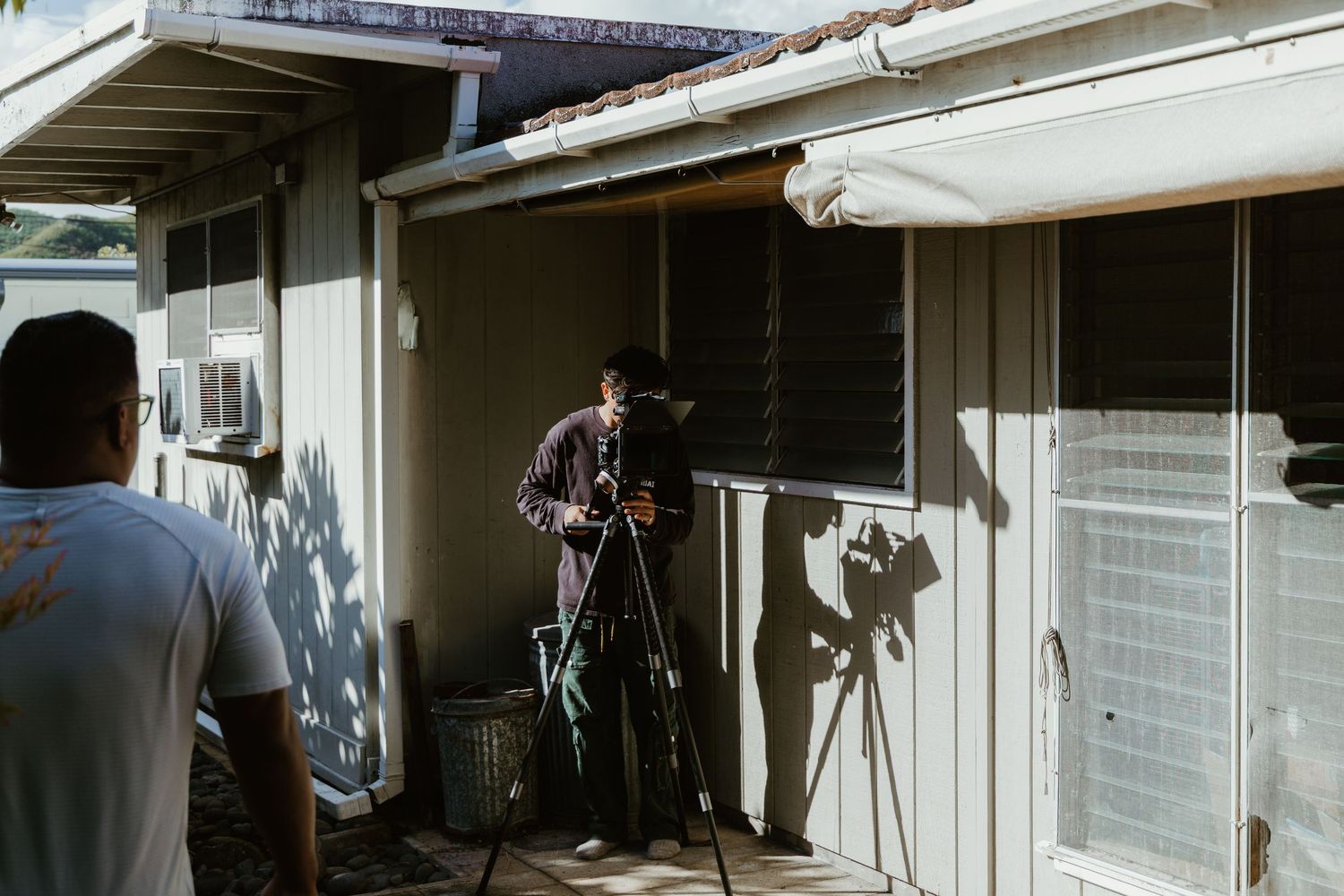 Filmmaker operating a camera on tripod outside a building, capturing footage in natural daylight.