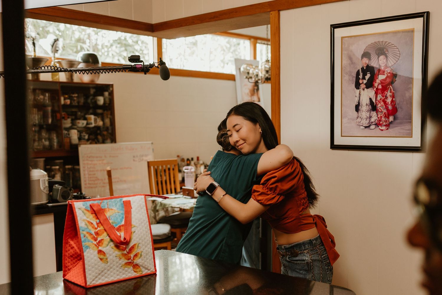 Two women sharing a warm hug indoors near a table with colorful gift bags and framed artwork on the wall.