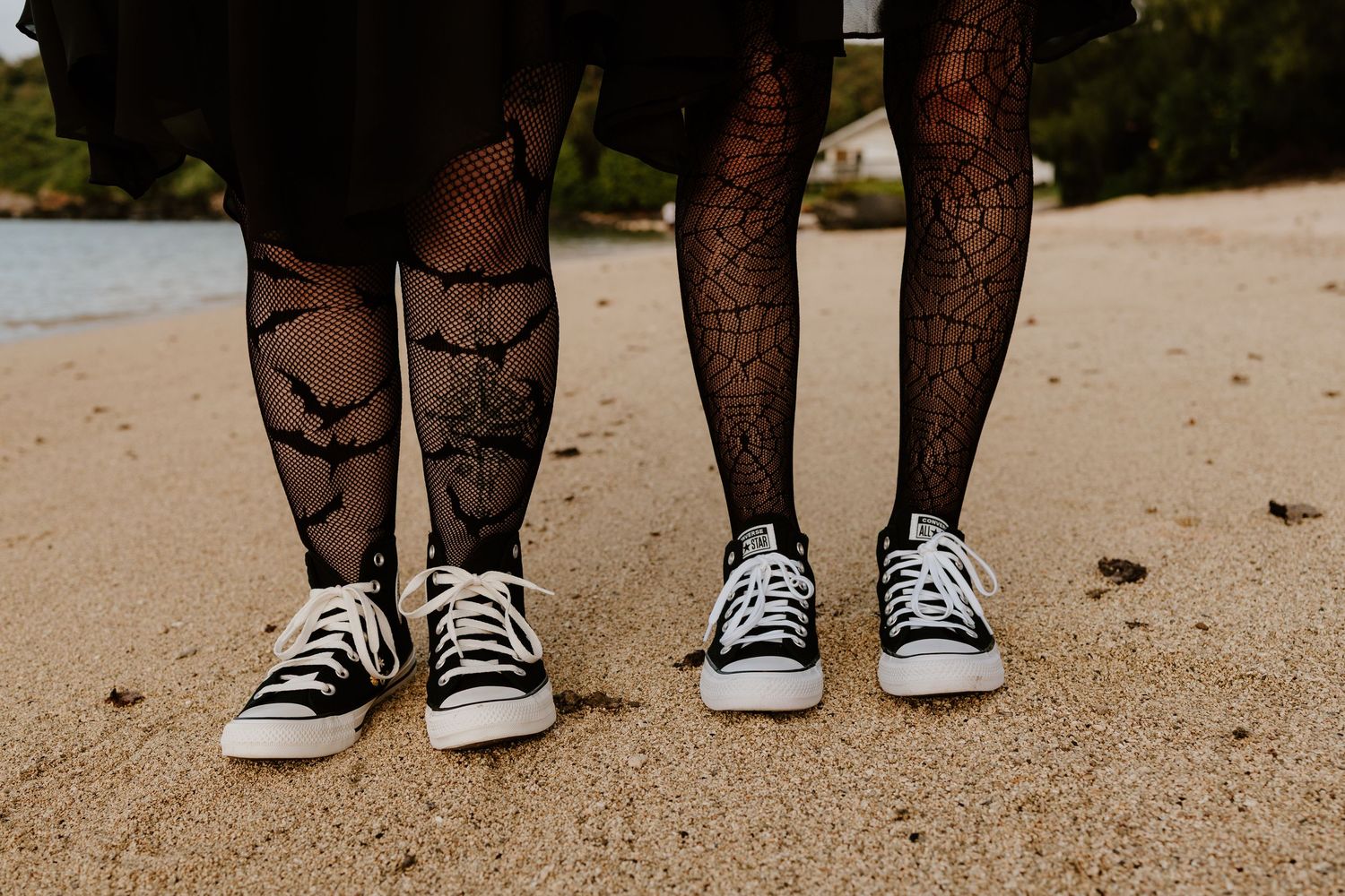 Two people stand on a sandy beach wearing black spiderweb-patterned tights and black-and-white Converse high-top sneakers.