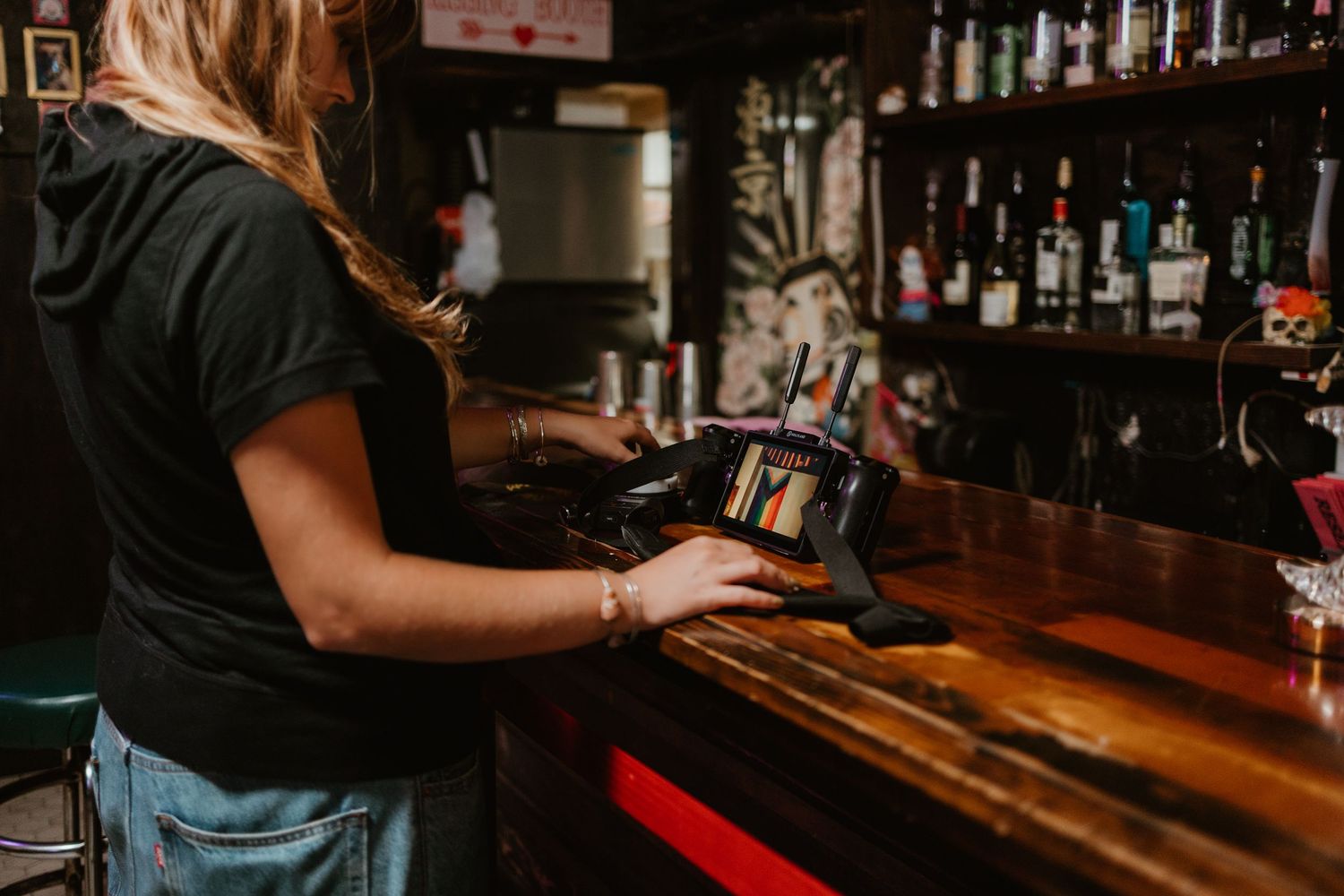 Female bartender using a POS system at a wooden bar counter with liquor bottles displayed on shelves behind her.