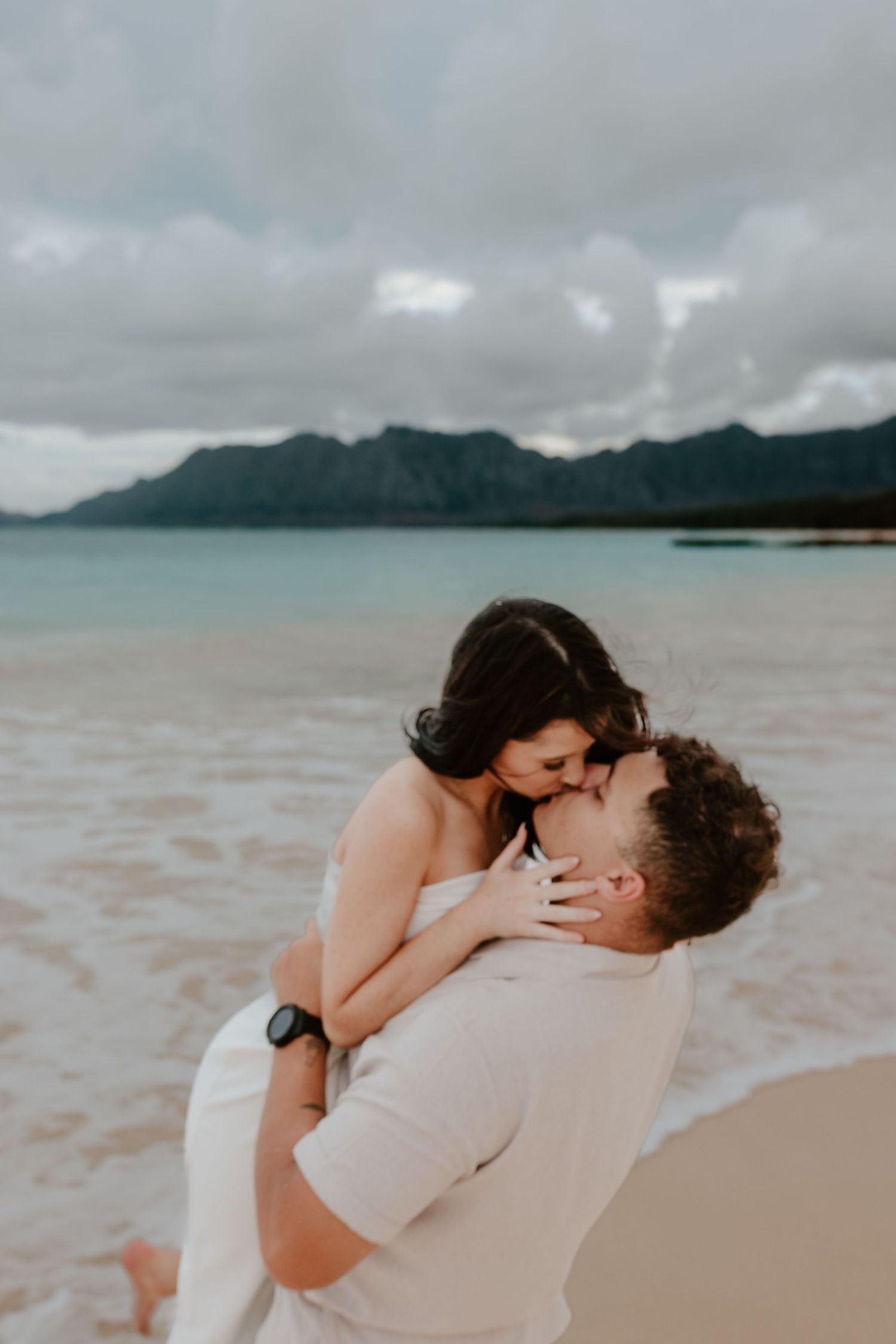 Man lifting woman in white dress as they share a kiss on a sandy beach with green mountains behind them.