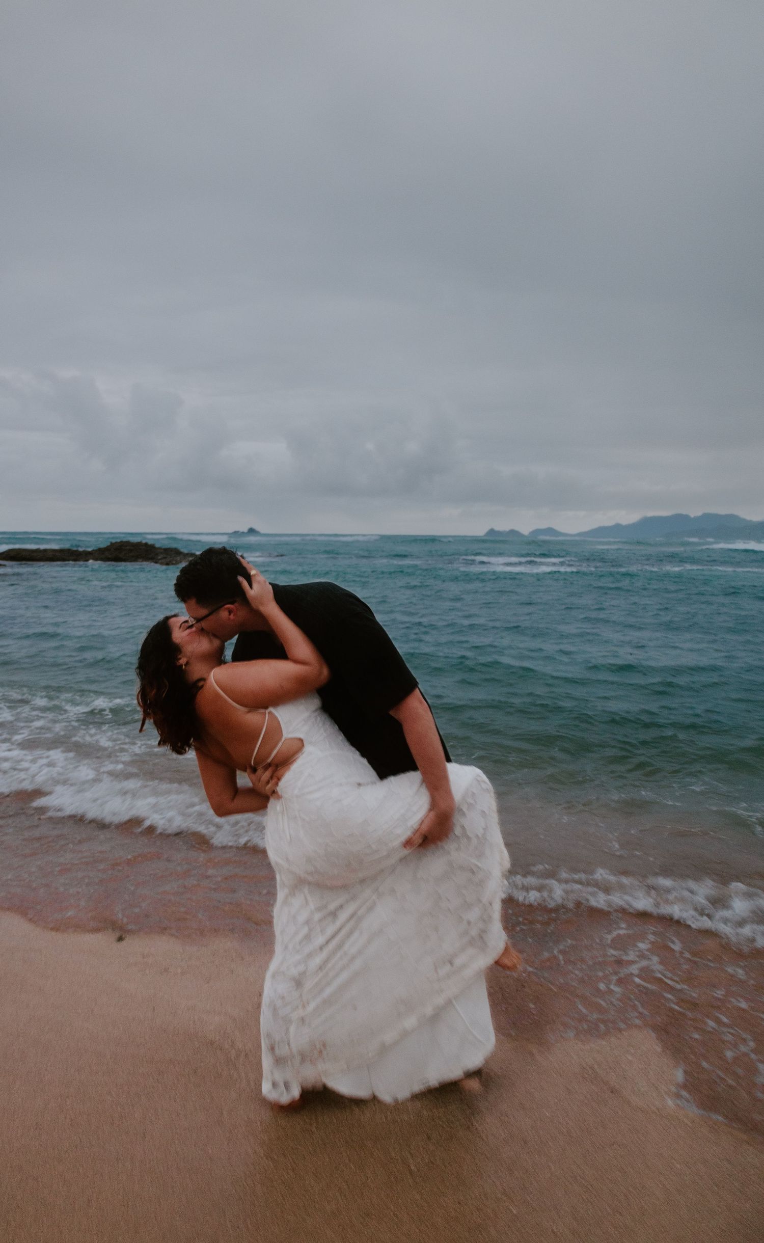 A groom dips his bride for a passionate kiss at the ocean shoreline under a dramatic cloudy sky.