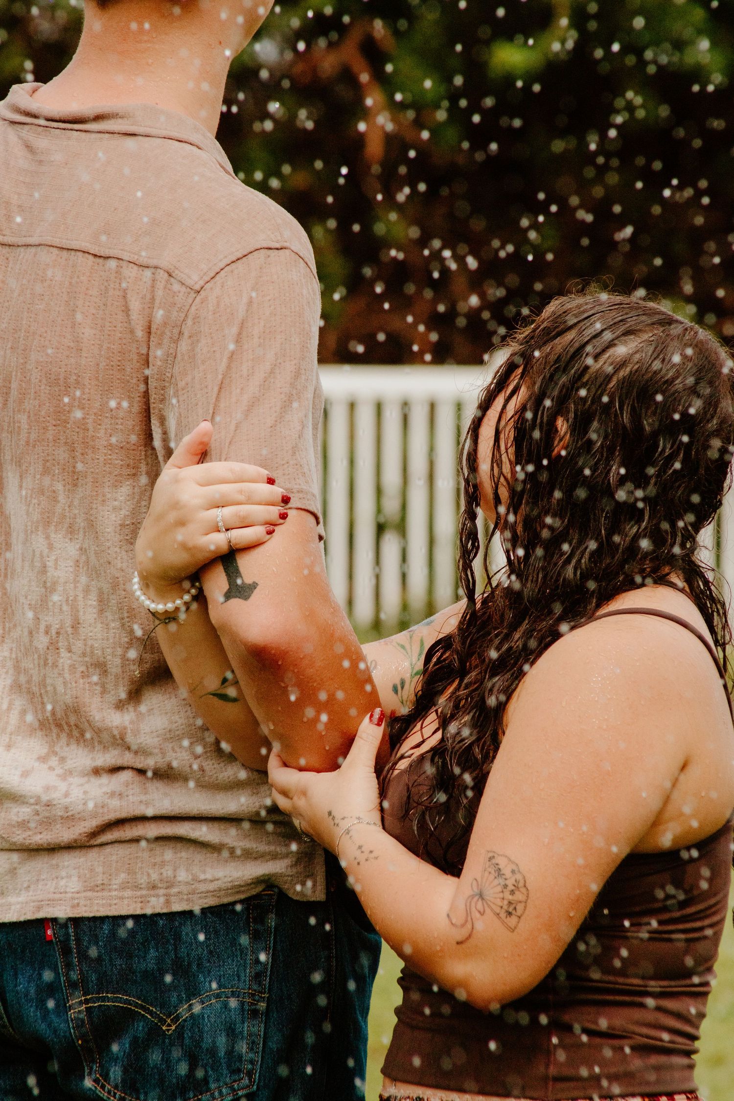 Couple standing close together in the rain, woman with tattoos and wet hair holding man's arm outdoors.