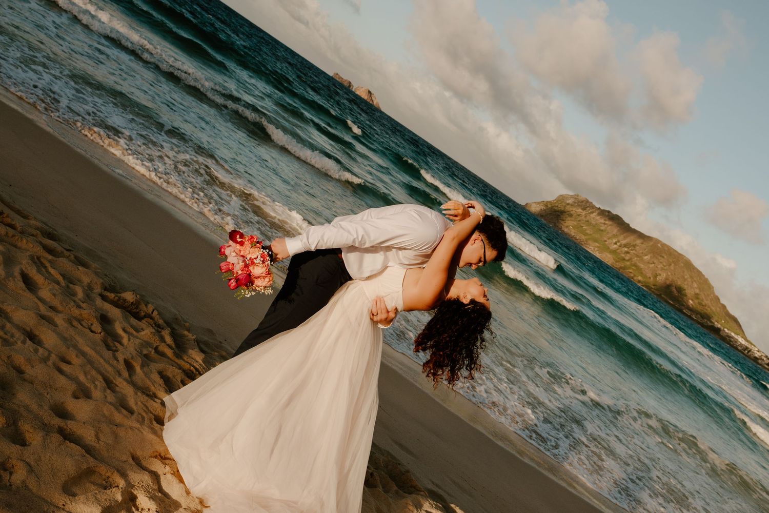 Groom dips bride in white wedding dress on sandy Hawaii beach with turquoise ocean waves in background.