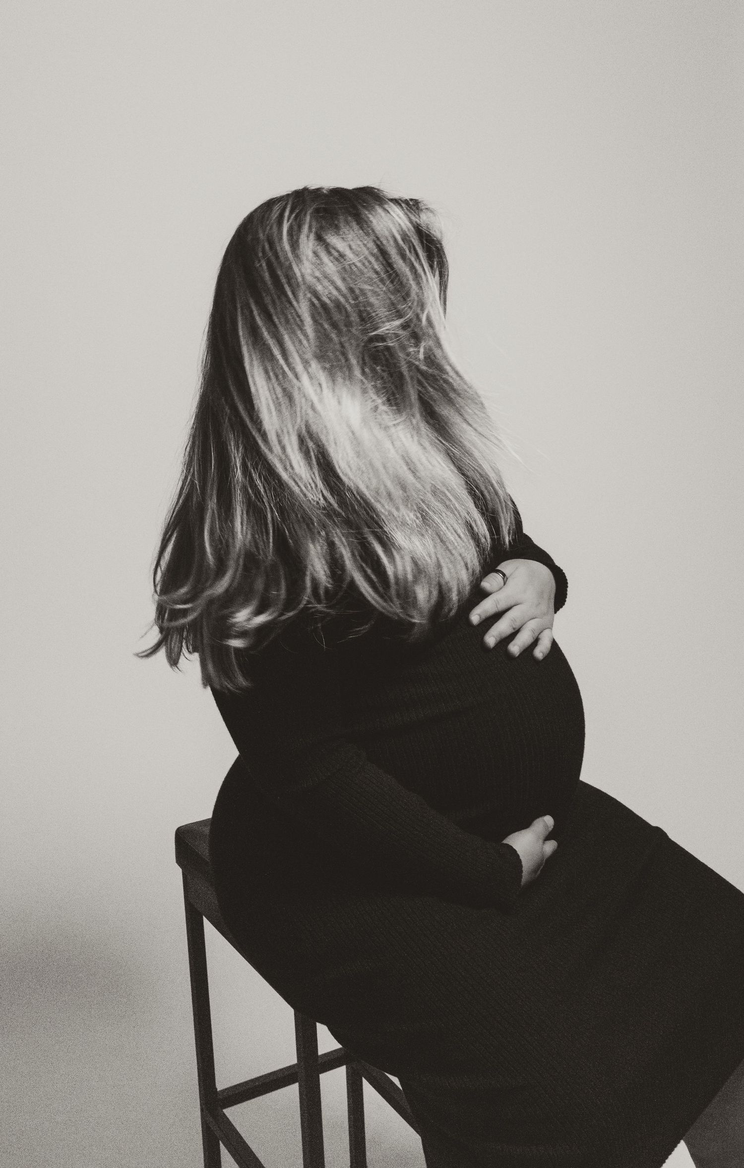 Black and white rear view of pregnant woman with long hair sitting on stool holding her bump.