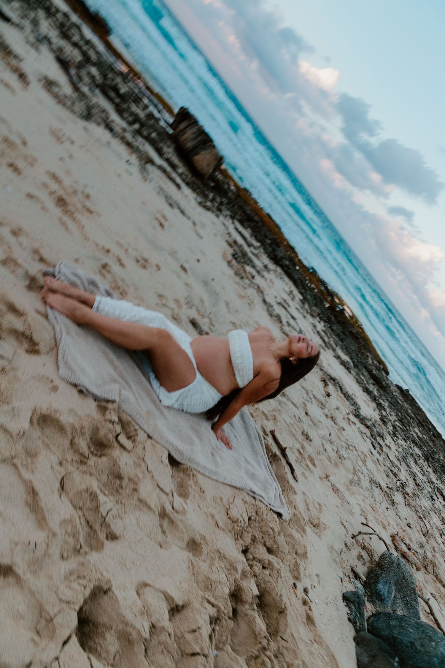 Pregnant woman in white crop top lying on sandy beach at sunset with turquoise ocean waves in background.