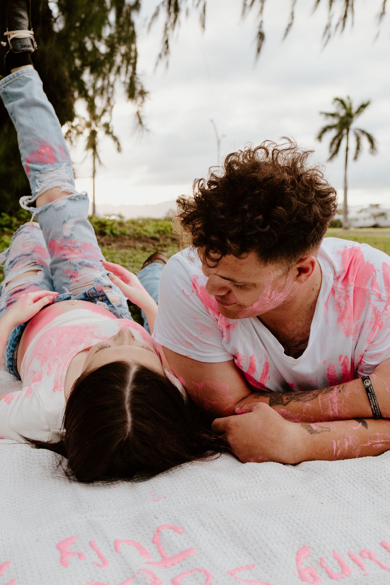 Couple covered in pink paint lying together on blanket smiling at each other outdoors.