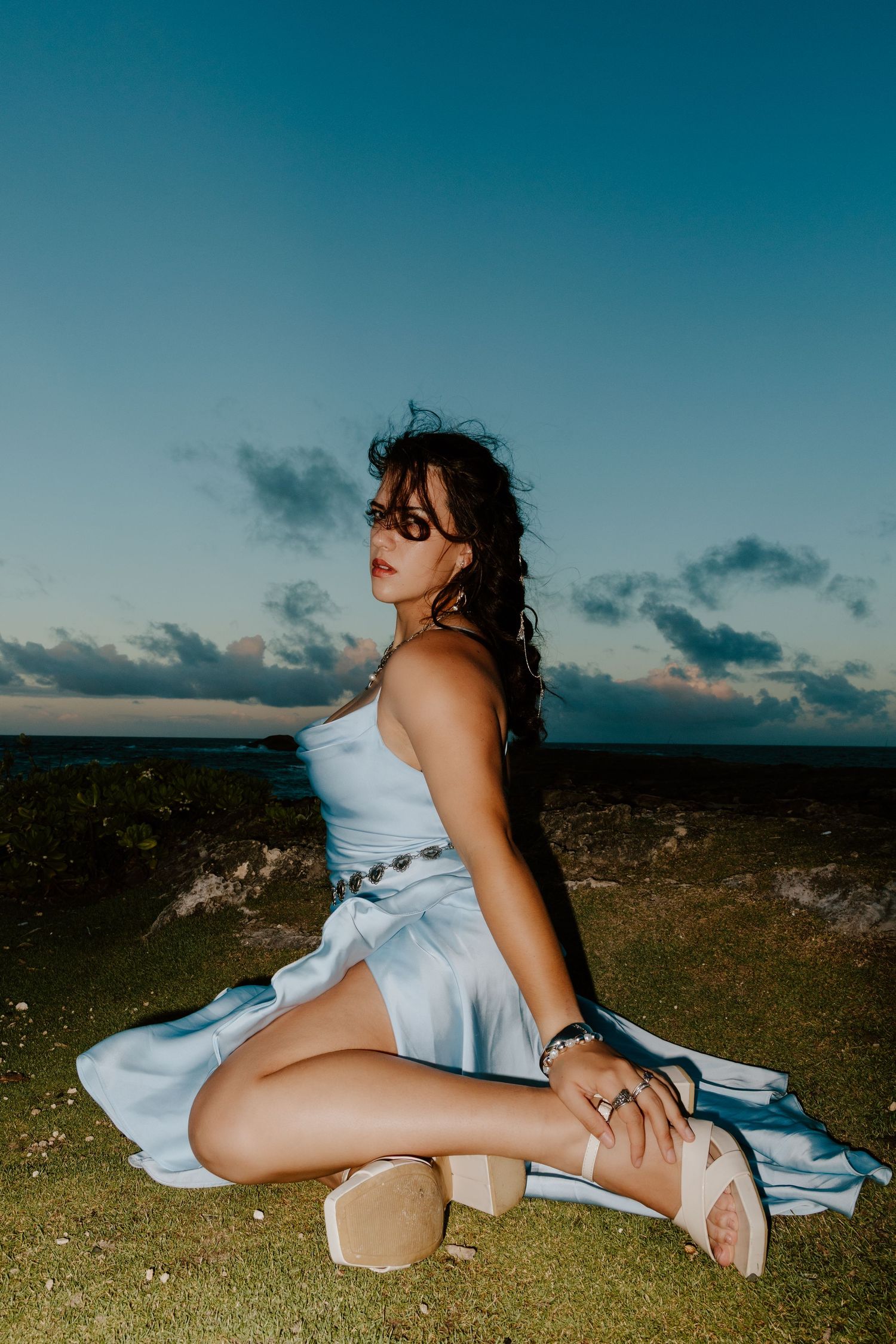 Woman in light blue satin dress sitting on grass cliff at sunset with ocean view behind her.