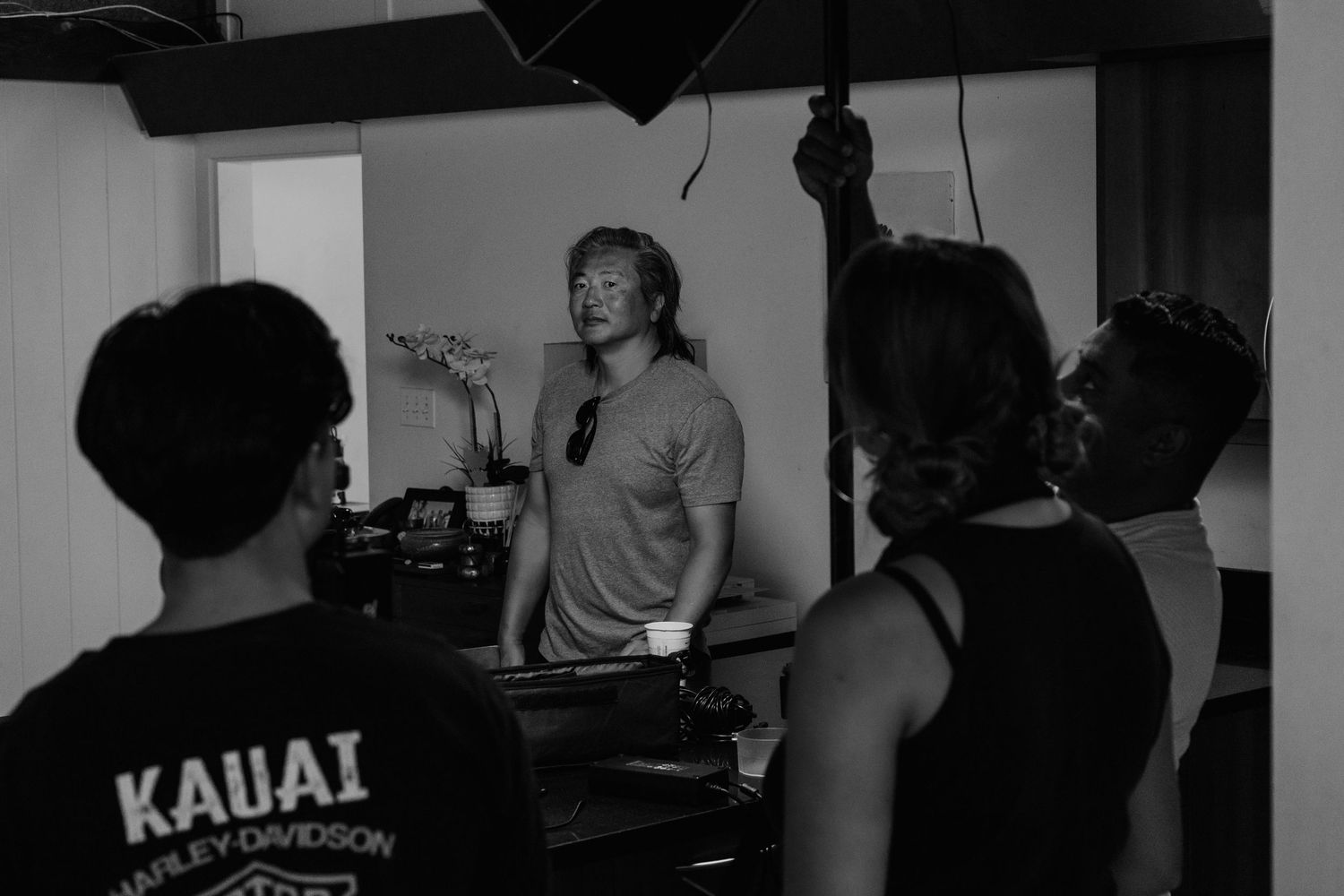 Black and white photo of people gathered in a kitchen, one person wearing a Kauai Harley-Davidson shirt.