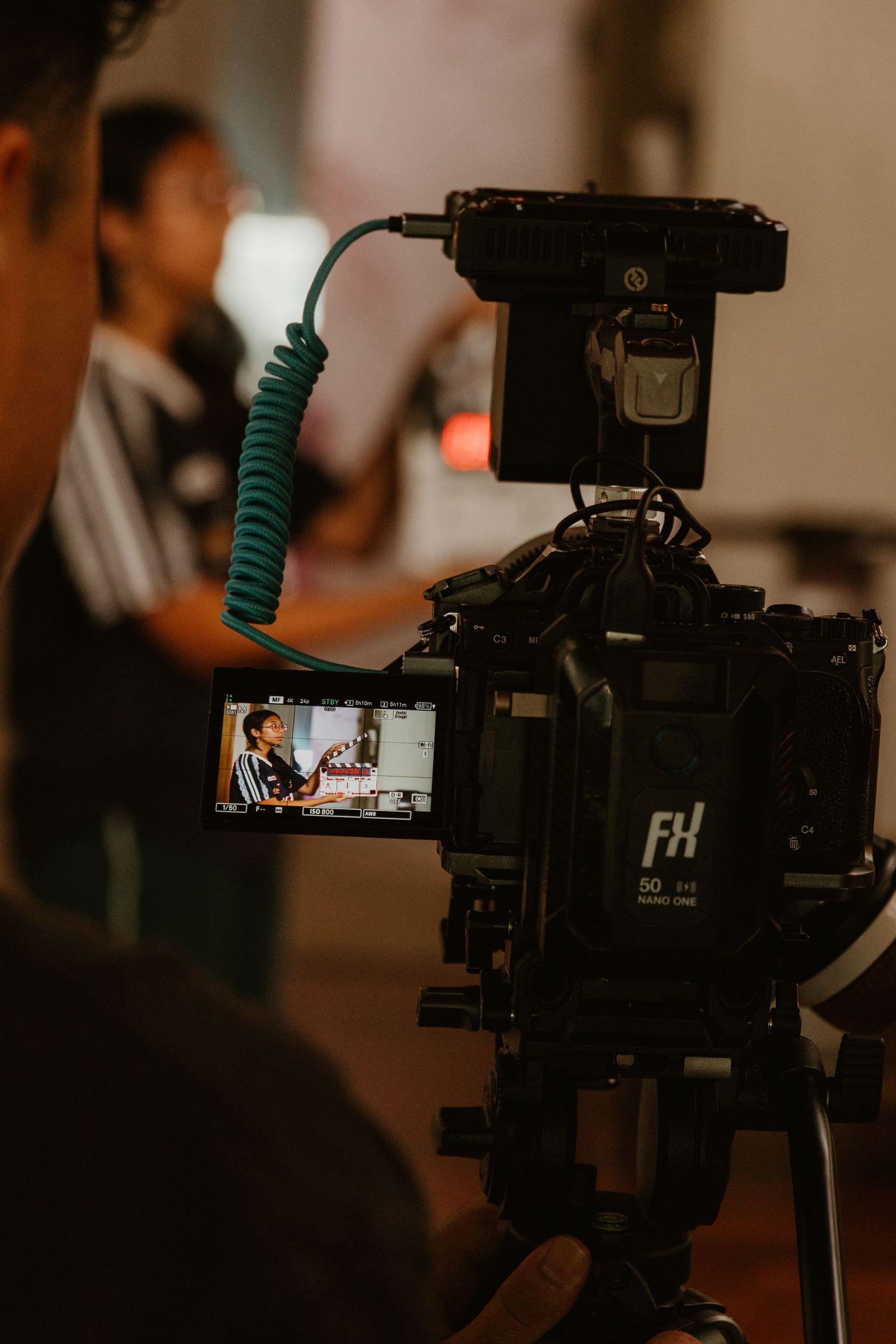 Professional video camera on tripod filming a person during a production shoot with crew visible in background.