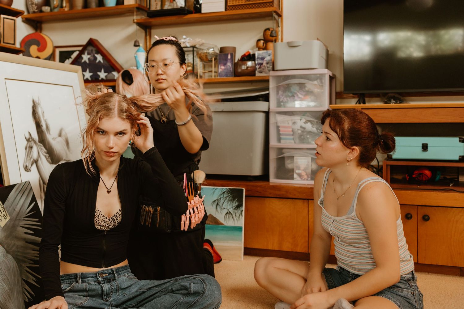 Three young women in a cozy room, one getting hair styled, sitting casually on the floor together.