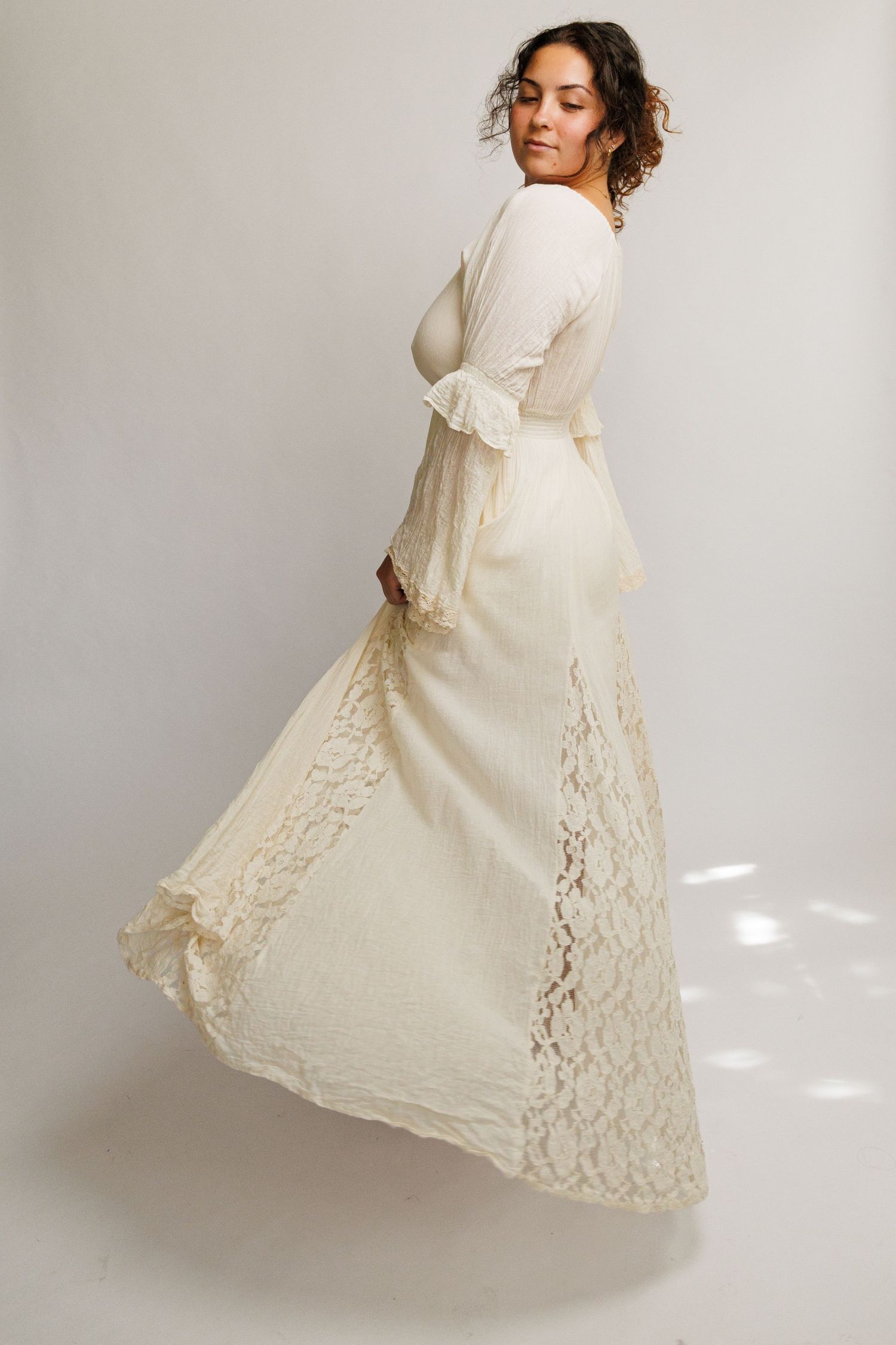 Woman in flowing ivory lace bridal gown with bell sleeves, twirling gracefully against a white background.