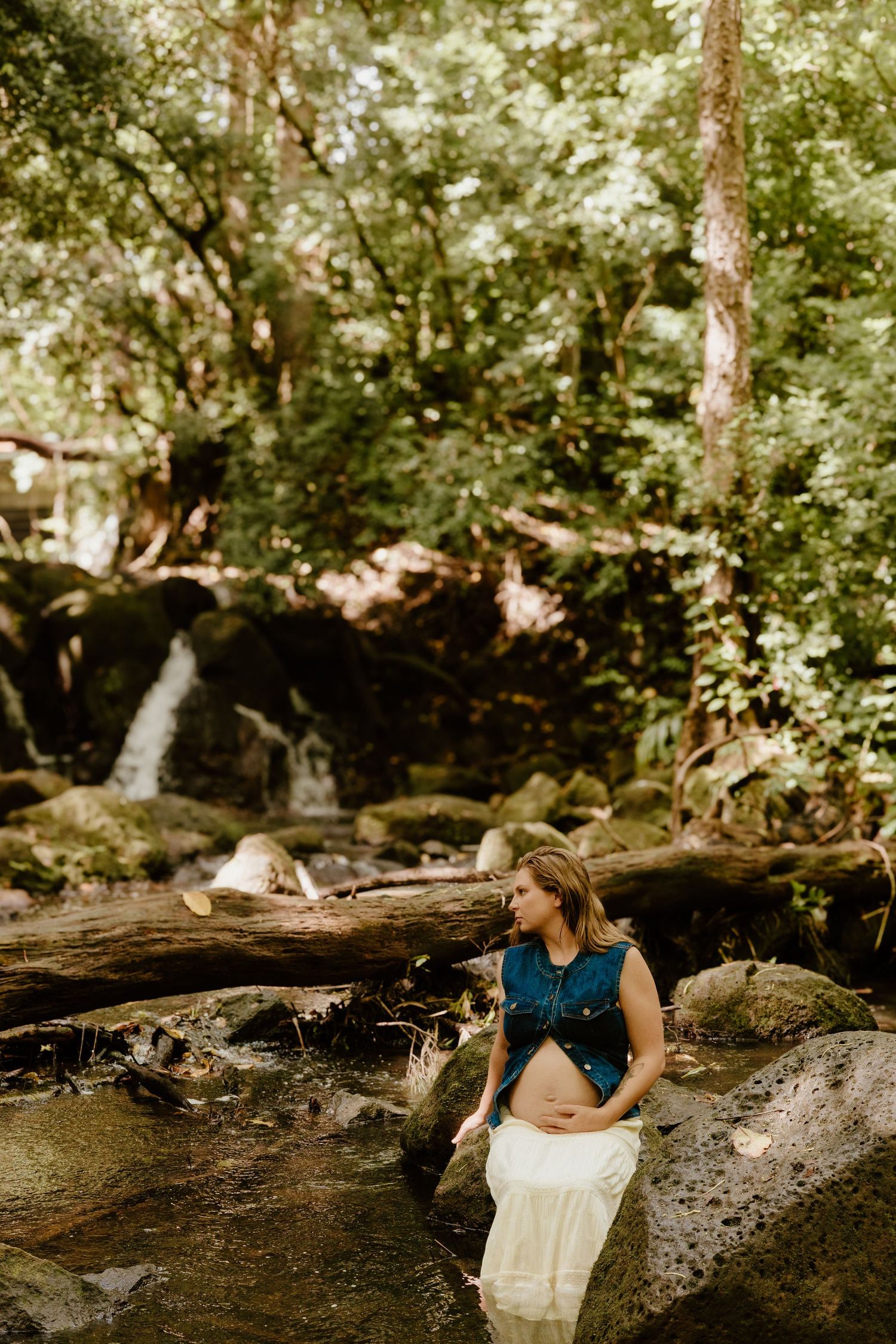 Pregnant woman in denim vest and white skirt standing in forest stream with waterfall background.