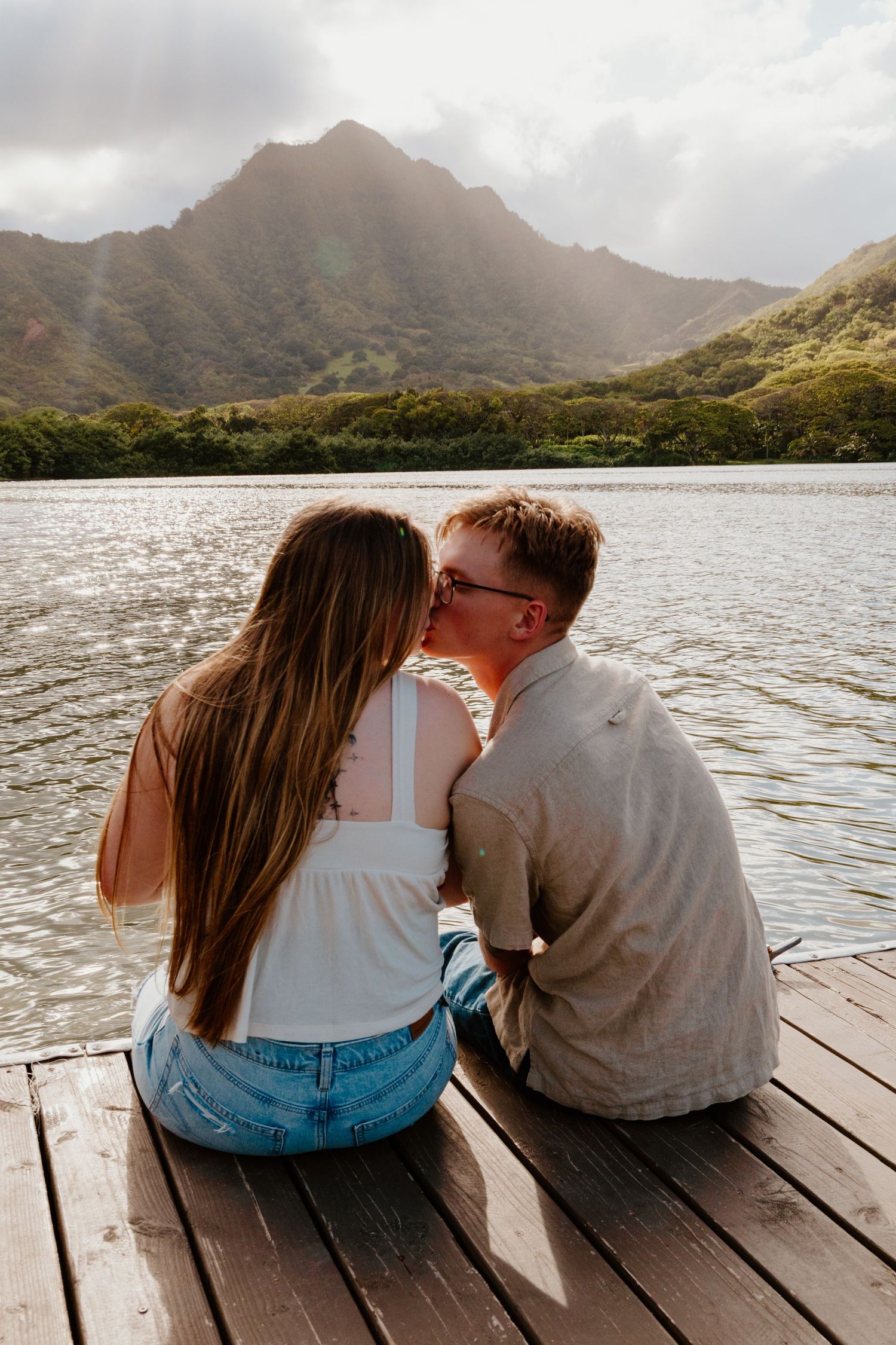 A couple shares a kiss while sitting on a wooden dock overlooking a stunning mountain lake.