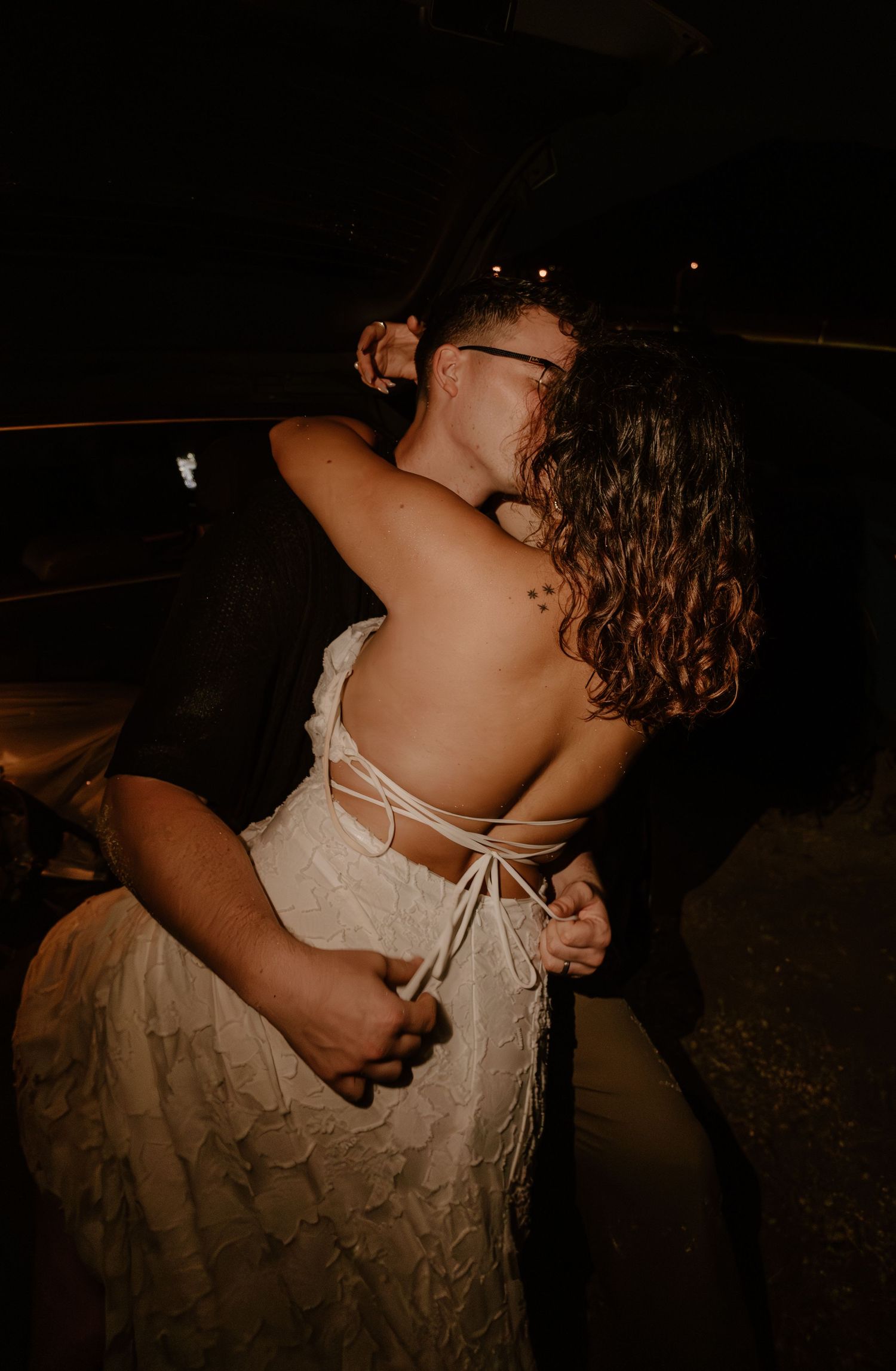 A couple shares an intimate embrace and kiss at night, bride in an open-back white lace dress.
