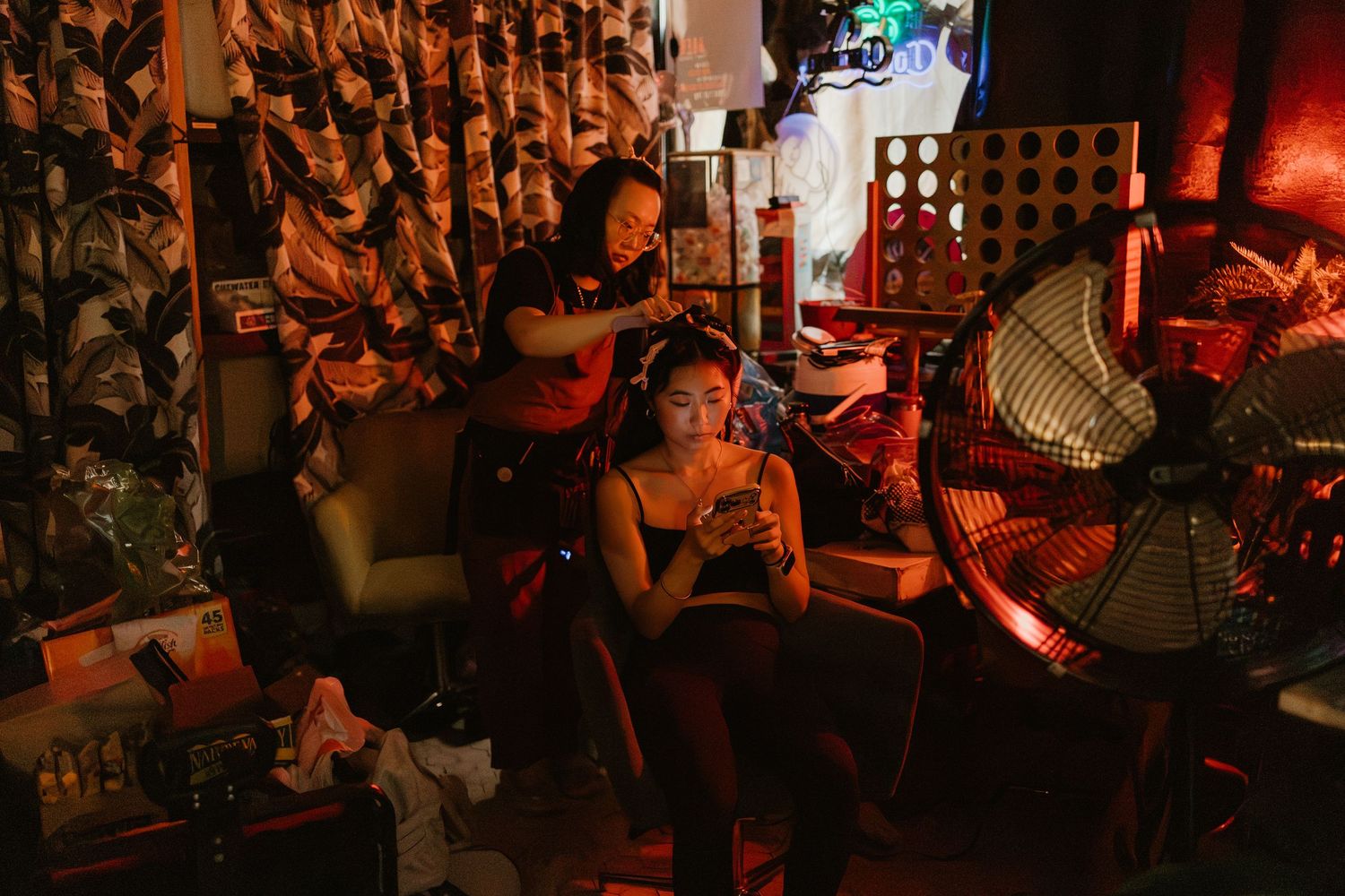 Two women in a dimly lit room with red lighting, one styling the other's hair near a fan.