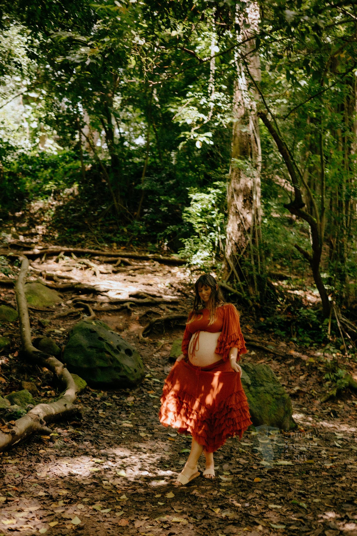 Pregnant woman in flowing rust dress walking through sunlit woodland forest path.