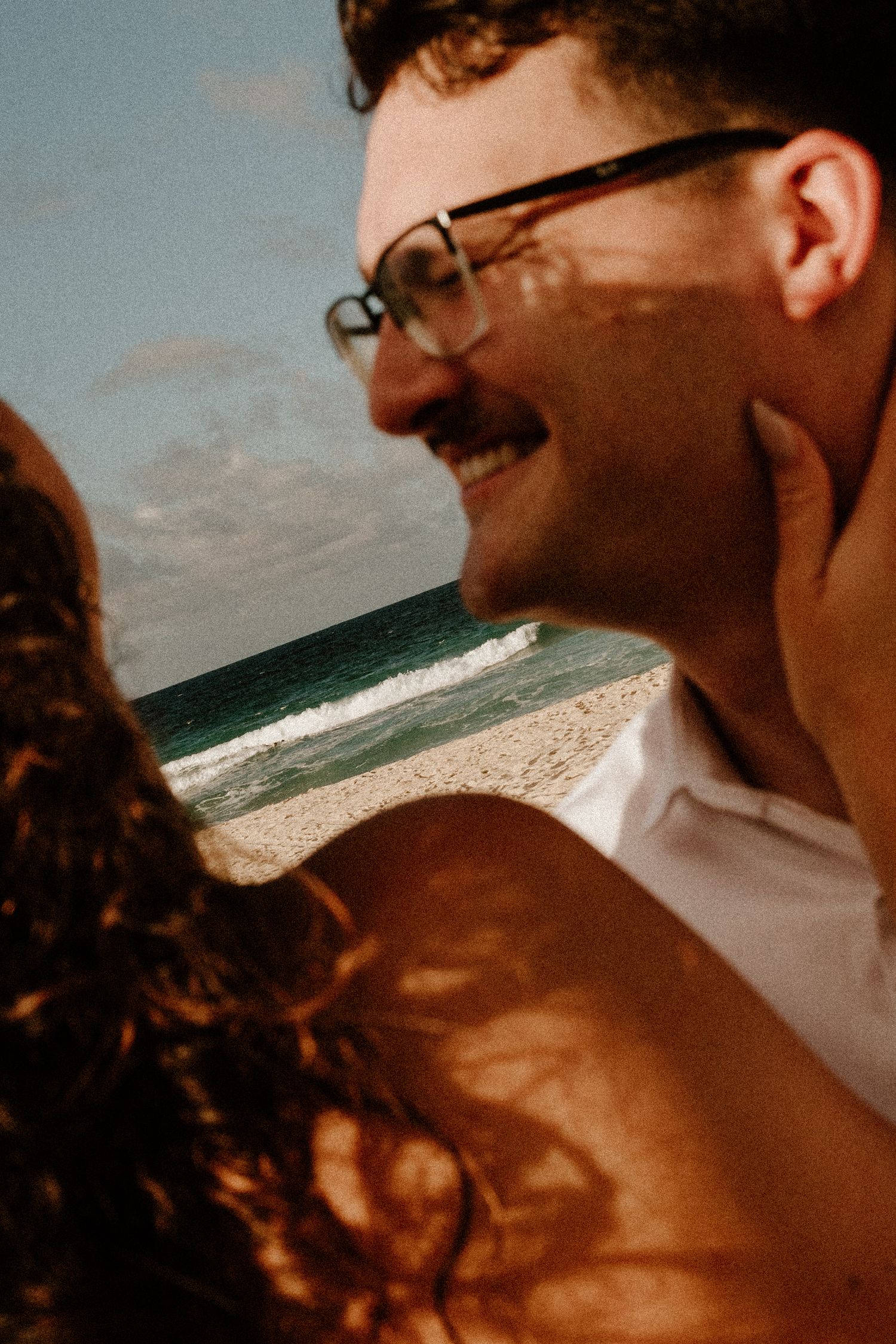 A smiling man with glasses and a mustache embraces a woman with curly red hair on a sunny beach.