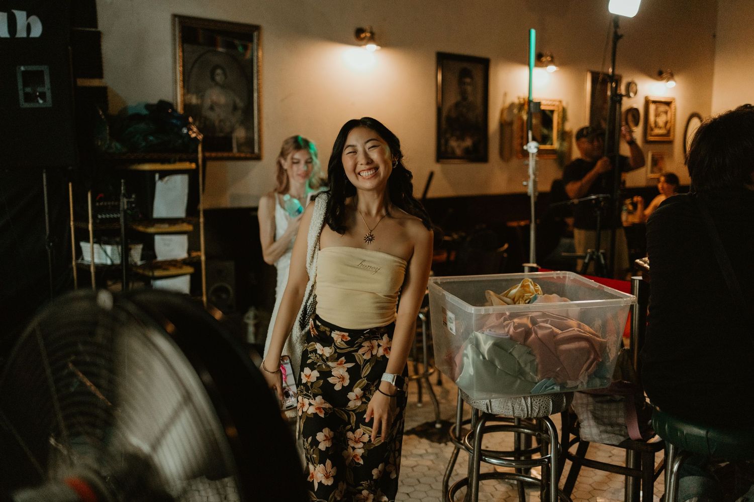 Smiling young woman in floral pants and tube top stands in a warmly lit tattoo shop or bar.